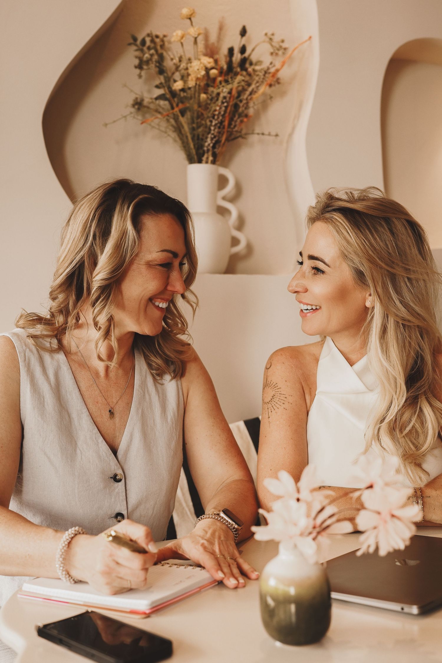 Two blonde women smiling and laughing together at a table with notebooks, flowers, and a laptop in a bright modern cafe.