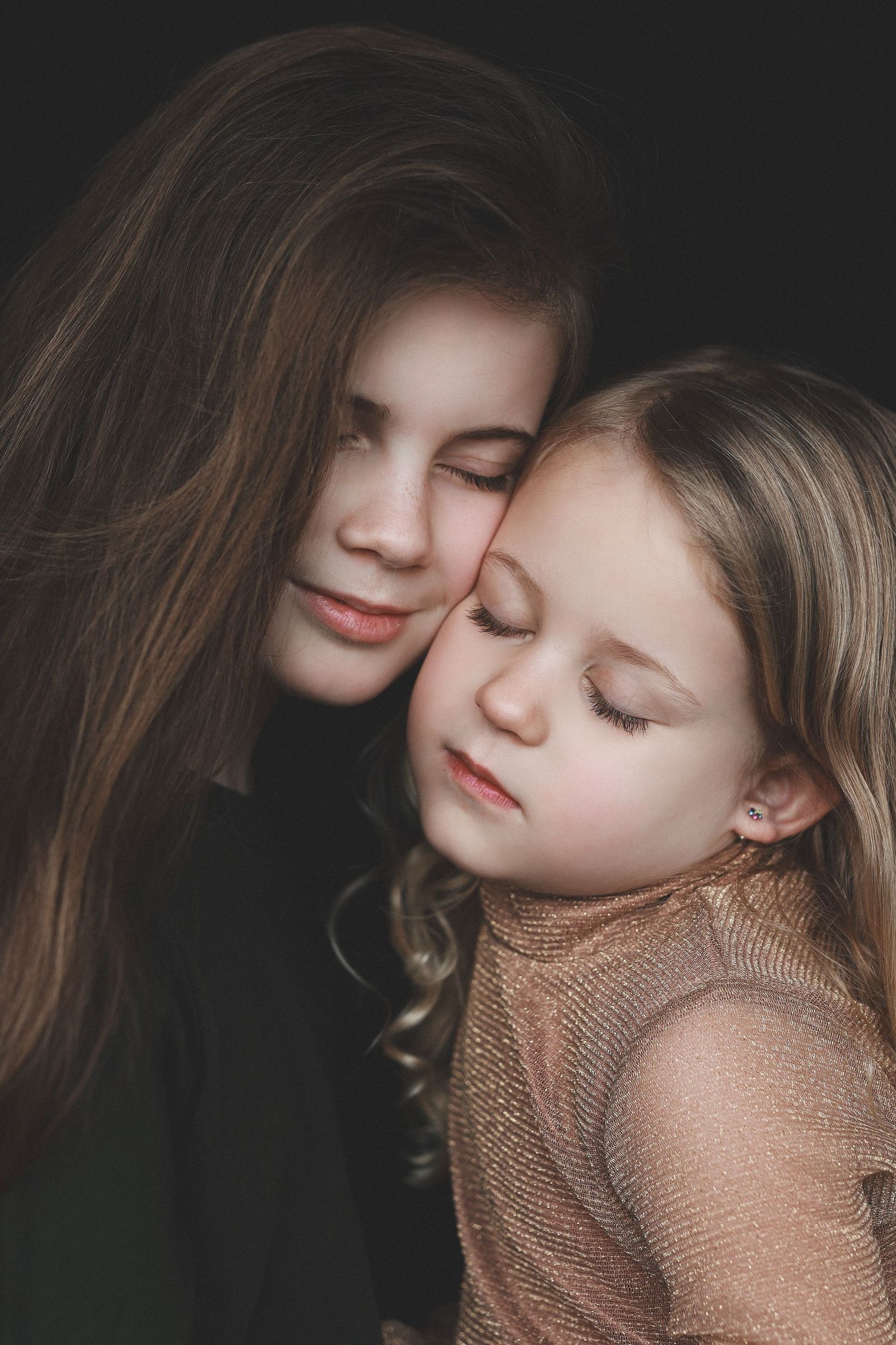 Two young girls with closed eyes share a tender moment, leaning heads together against a dark background.
