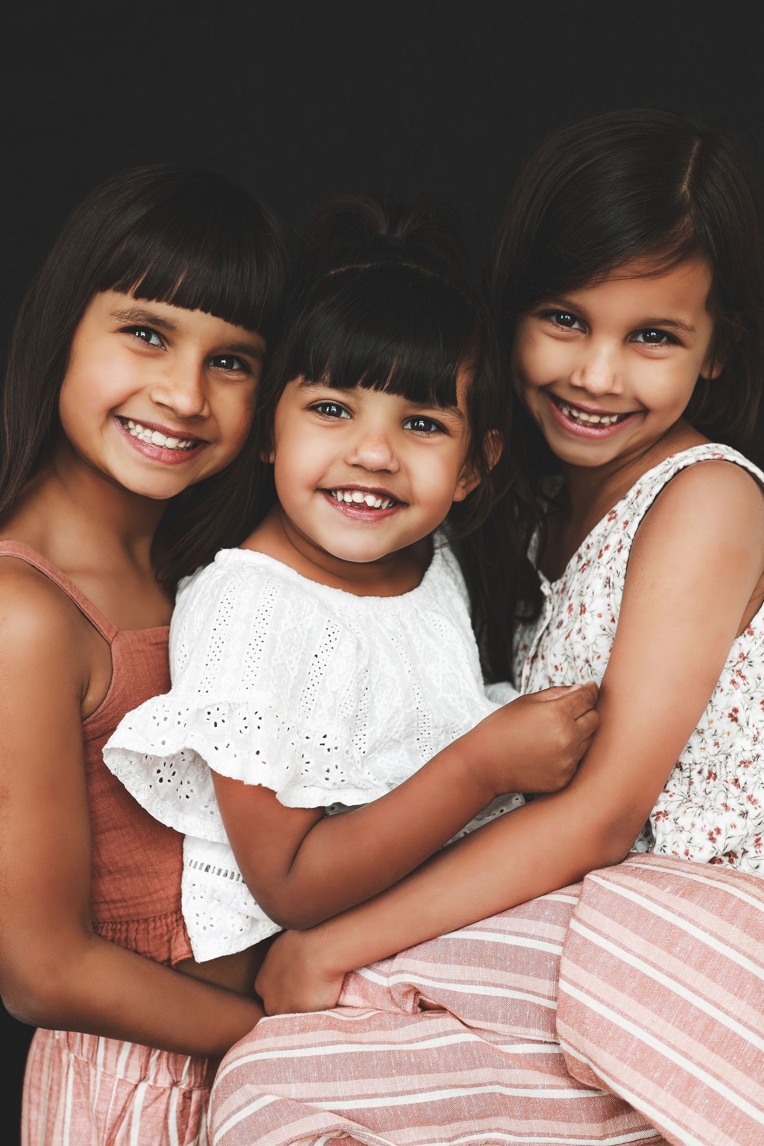 Three young sisters hugging and smiling in colorful dresses against dark background.