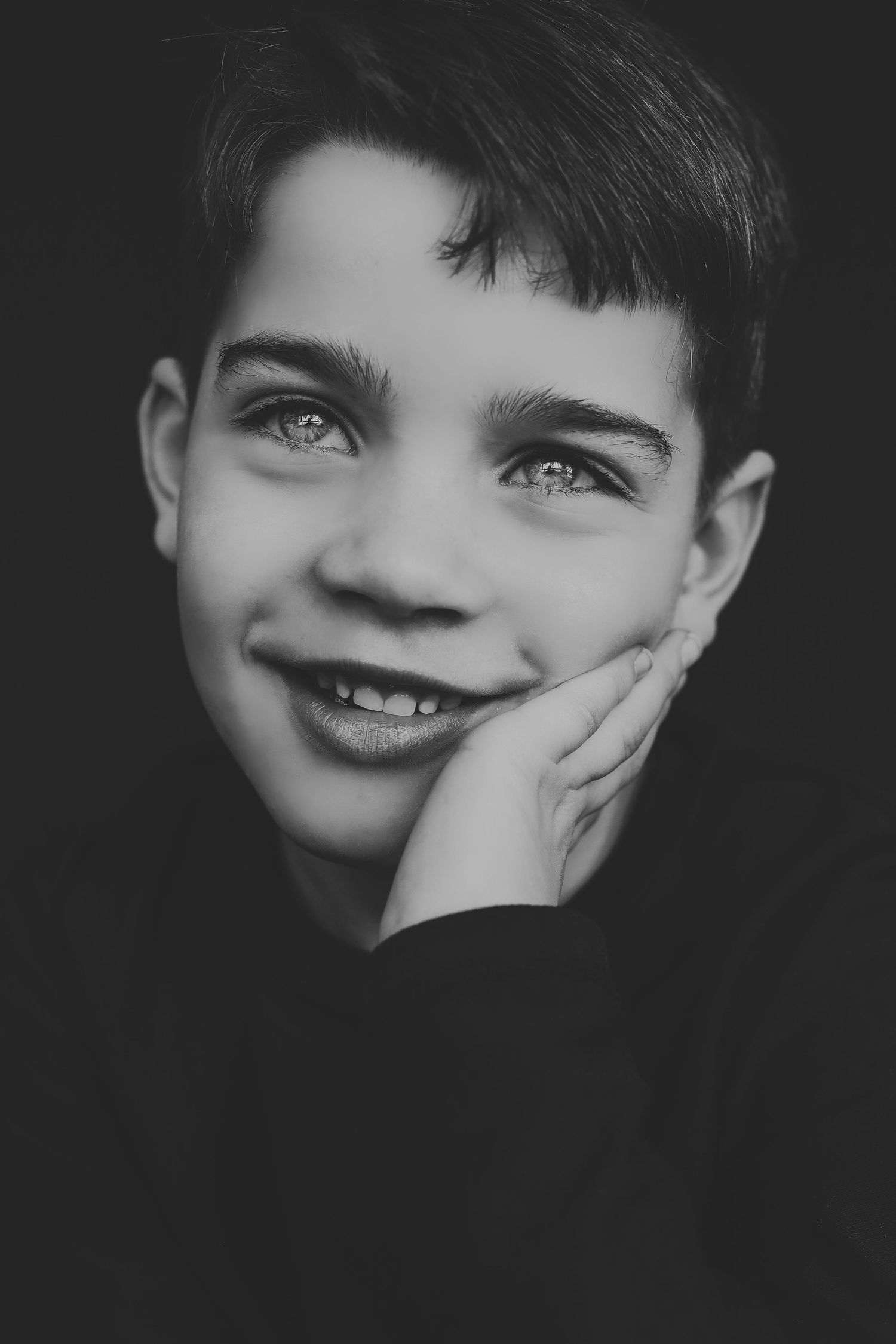Black and white portrait of a smiling young boy resting his chin on his hand against a dark background.