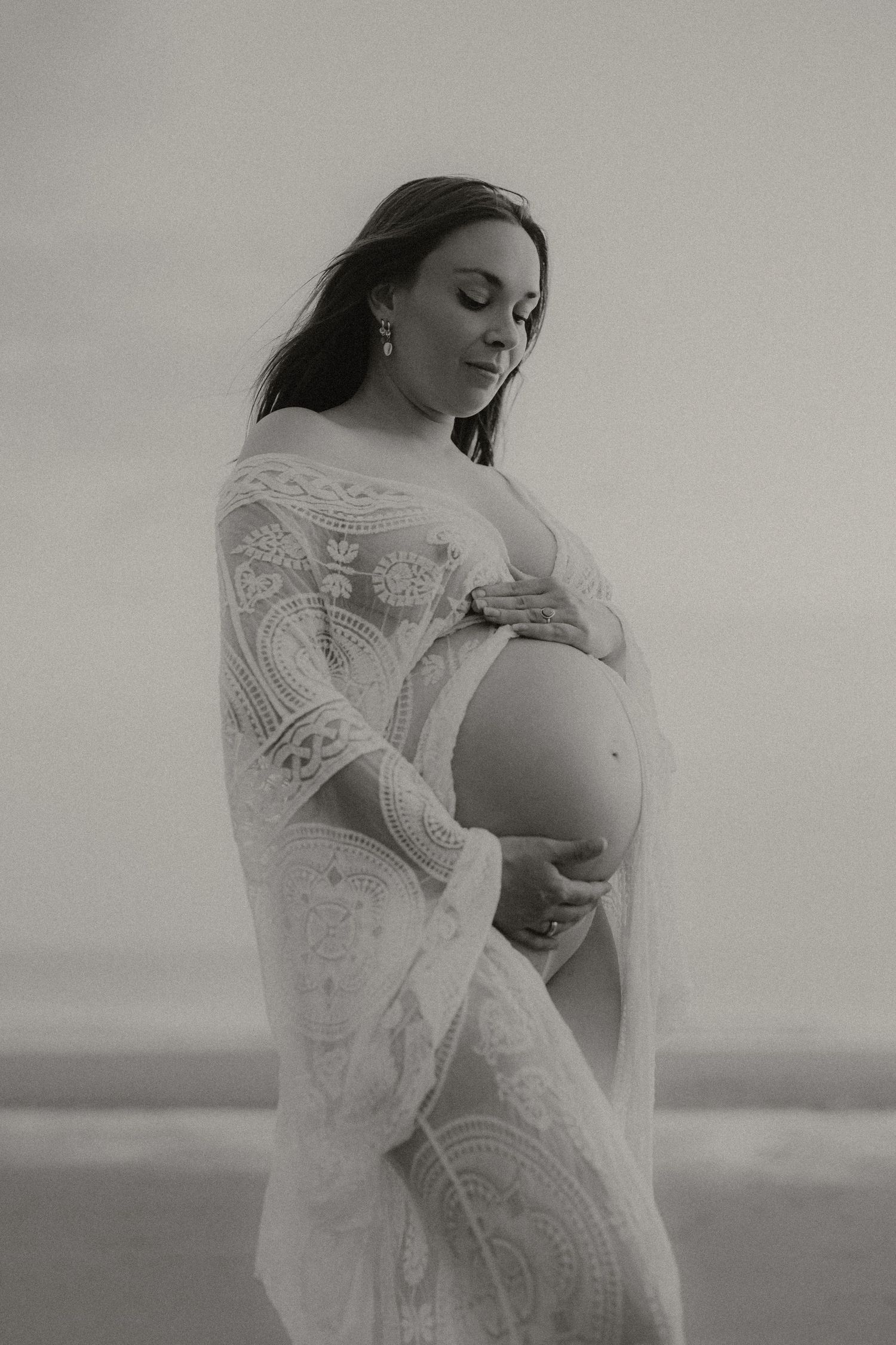 Black and white portrait of pregnant woman in lace dress cradling bare baby bump on beach.