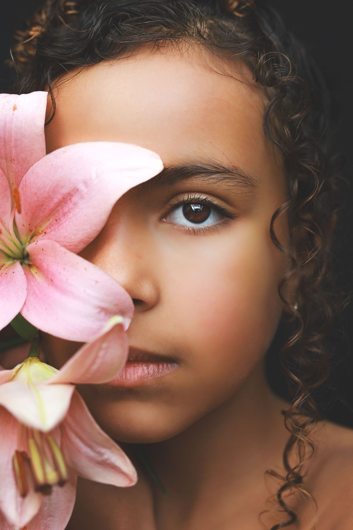 Close-up portrait of a young girl with curly hair and brown eyes partially hidden by pink lily.