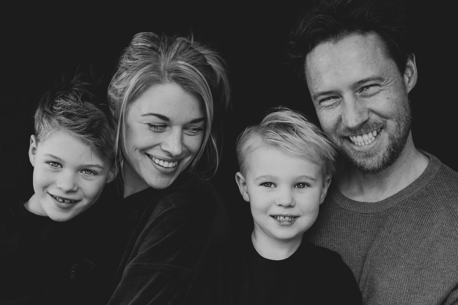 Black and white studio portrait of smiling family of four against dark background.