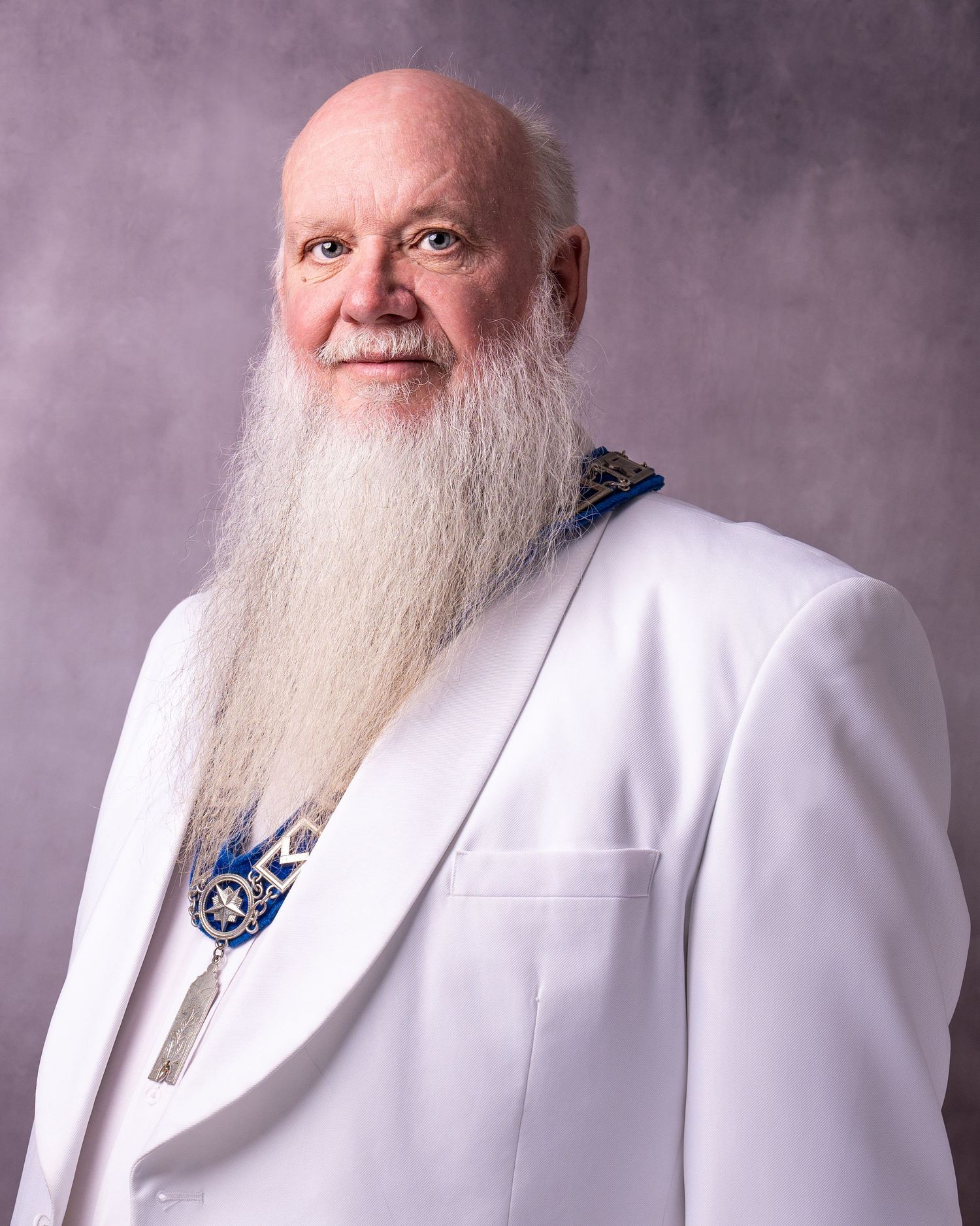Elderly man with long white beard wearing white formal jacket and blue decorative collar medallion.