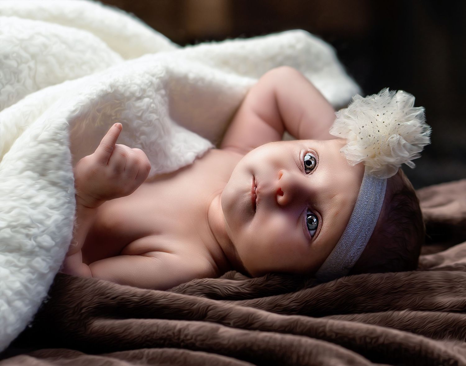 Newborn baby girl wearing a white floral headband, lying on a soft brown blanket wrapped in a white fluffy wrap.
