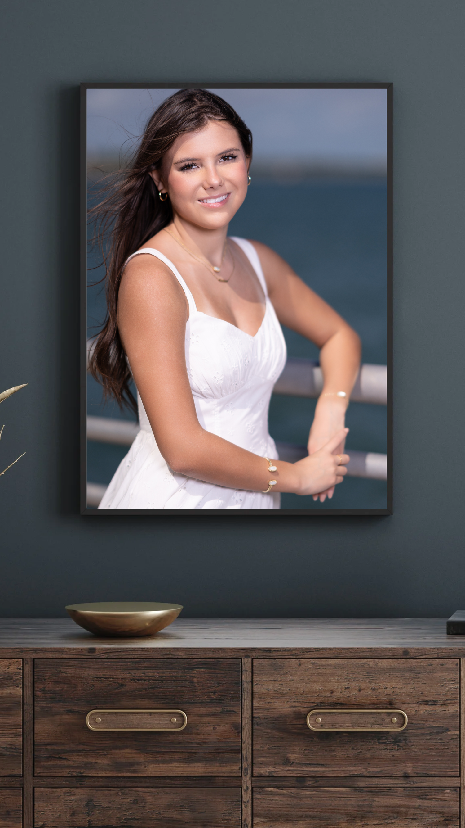 Young woman in white dress smiling outdoors near water, wearing gold jewelry, long dark hair blowing in breeze.