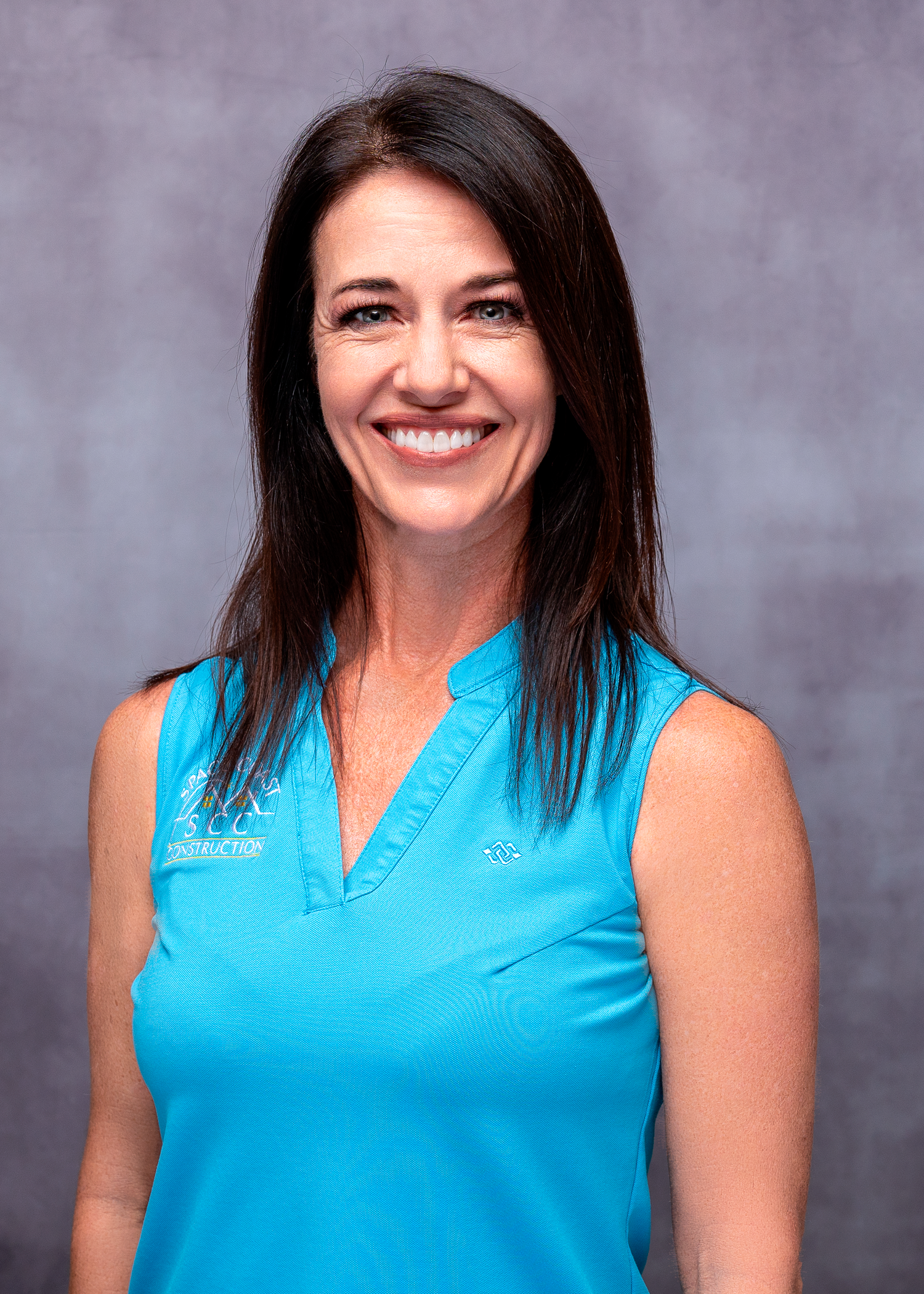 Smiling woman with dark hair wearing a turquoise sleeveless polo shirt against a gray background.