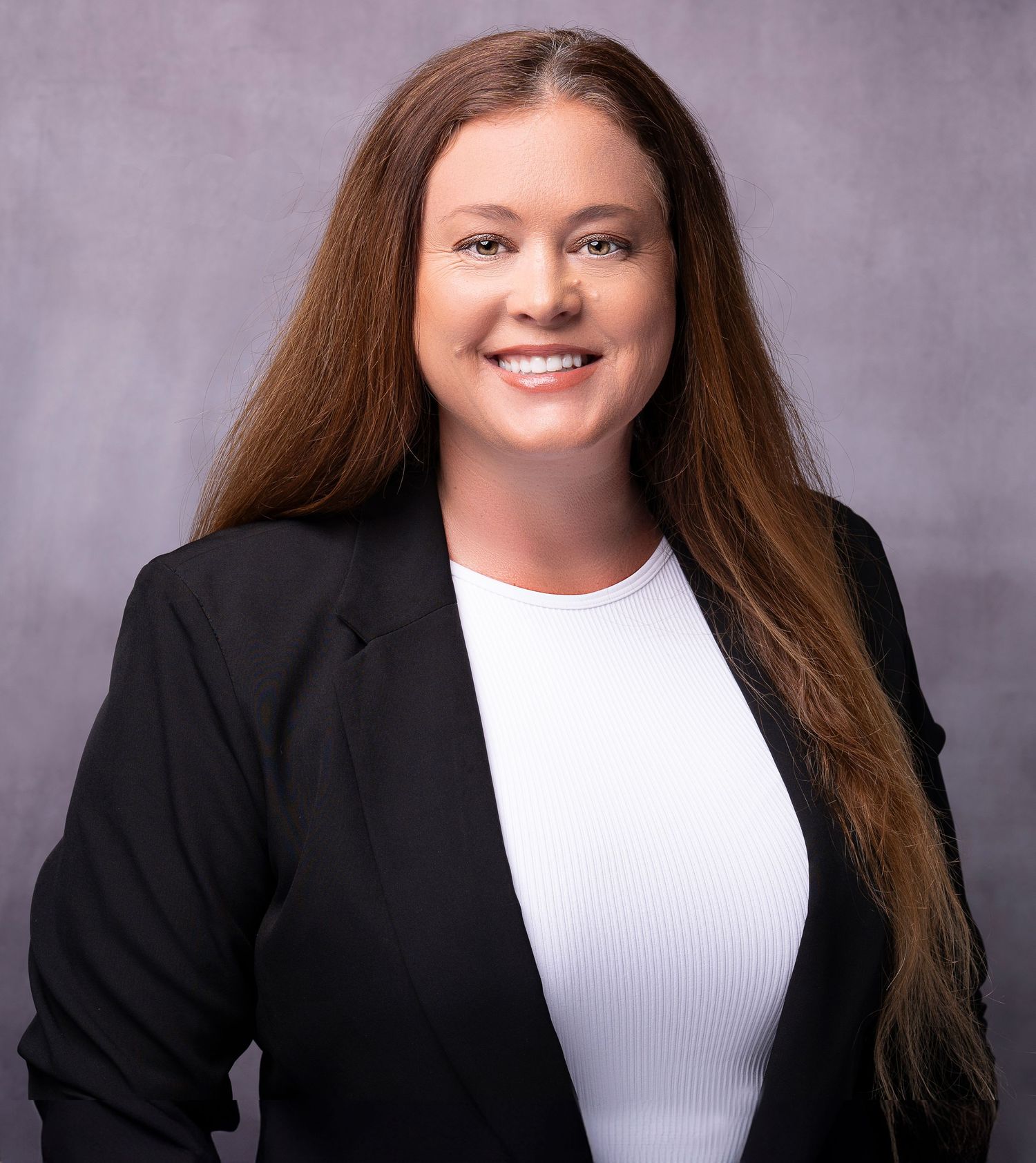 Professional headshot of a smiling woman with long brown hair wearing a black blazer and white top against a gray background.