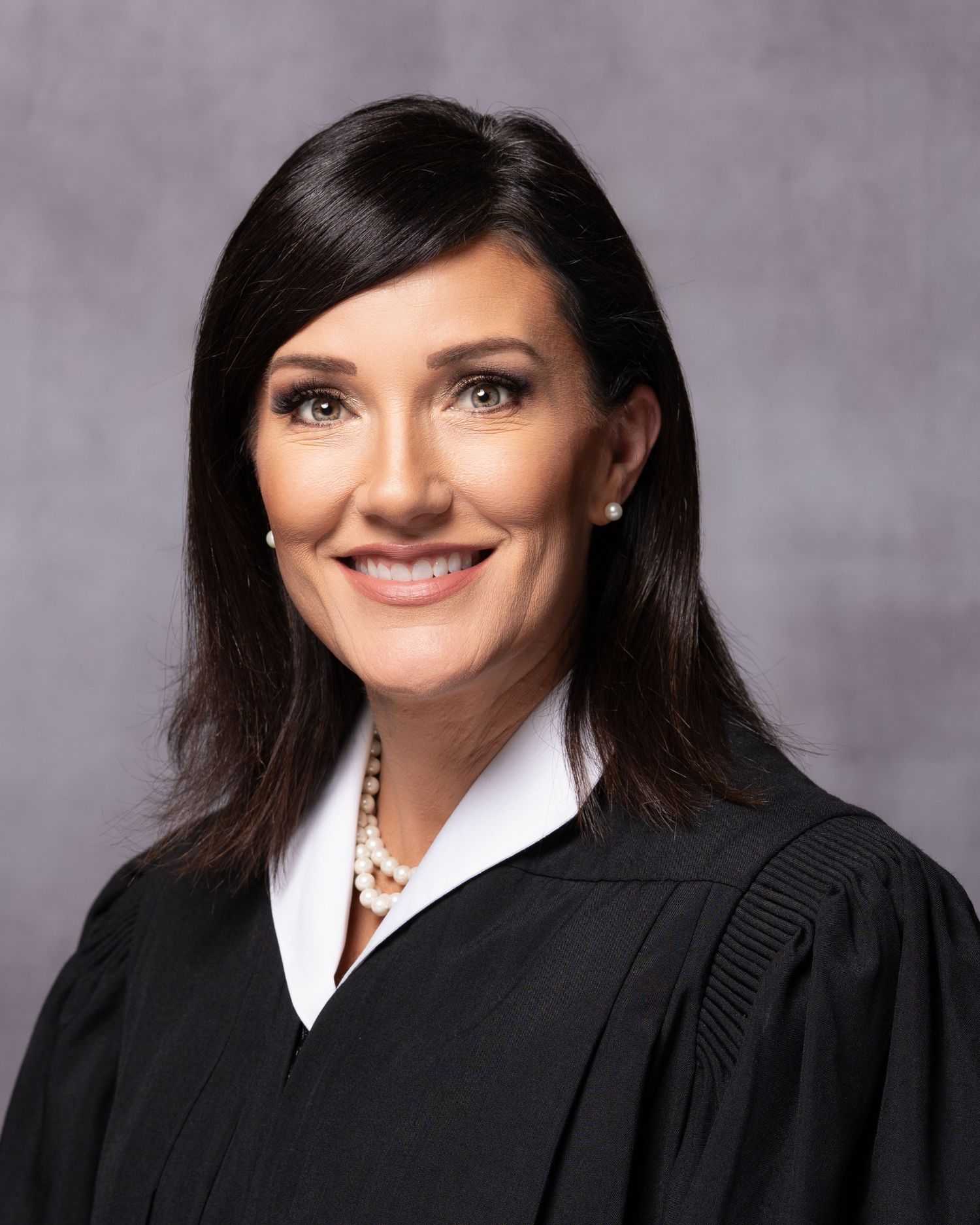 Professional portrait of a female judge wearing black robes and pearl jewelry, smiling confidently against a gray background.