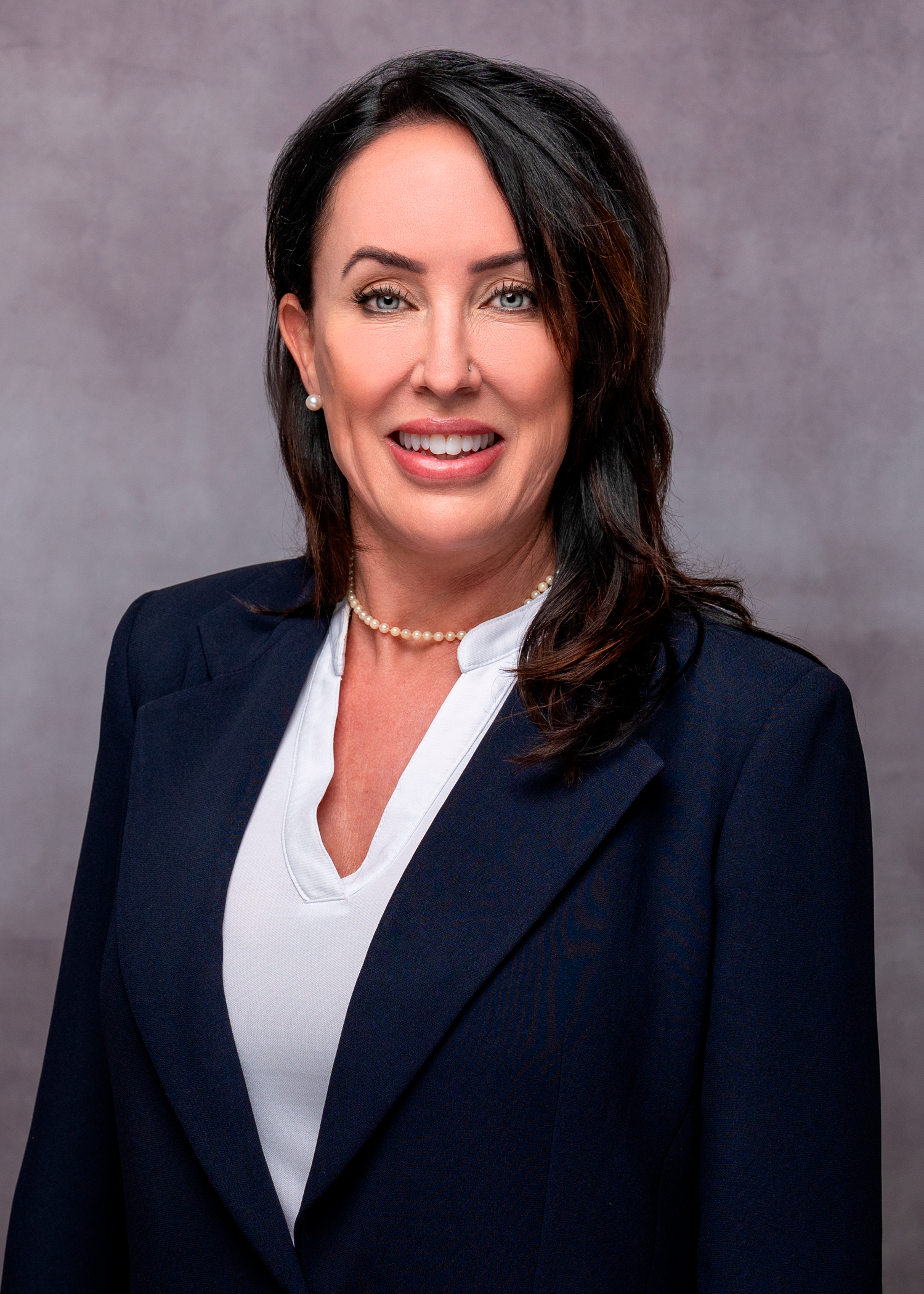 Professional headshot of a woman in a navy blazer and pearl necklace, smiling confidently against a gray background.