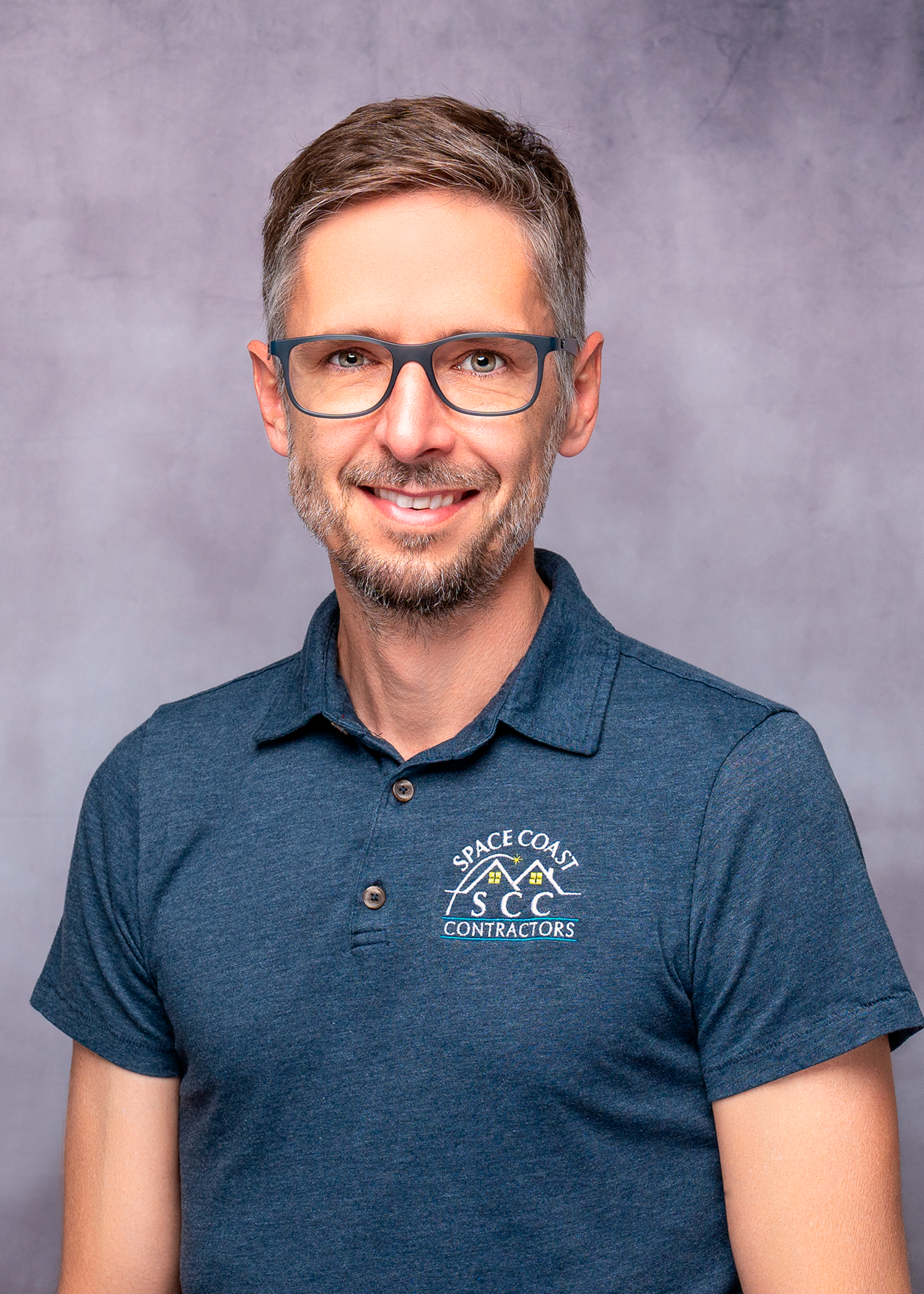 Professional headshot of a smiling man wearing glasses and a Space Coast Contractors polo shirt against a gray background.