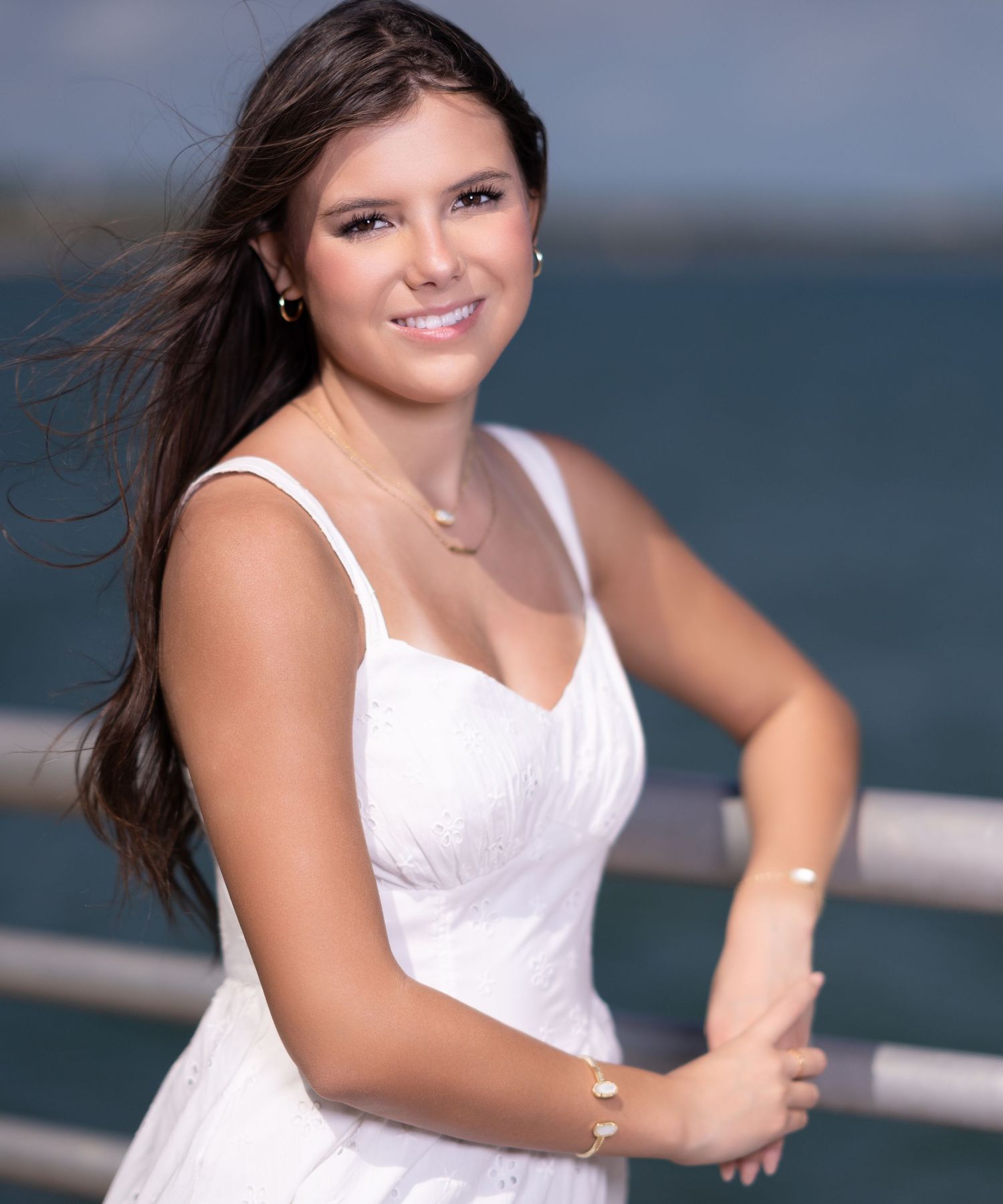 Young woman in white dress smiling outdoors near waterfront railing on a sunny day.