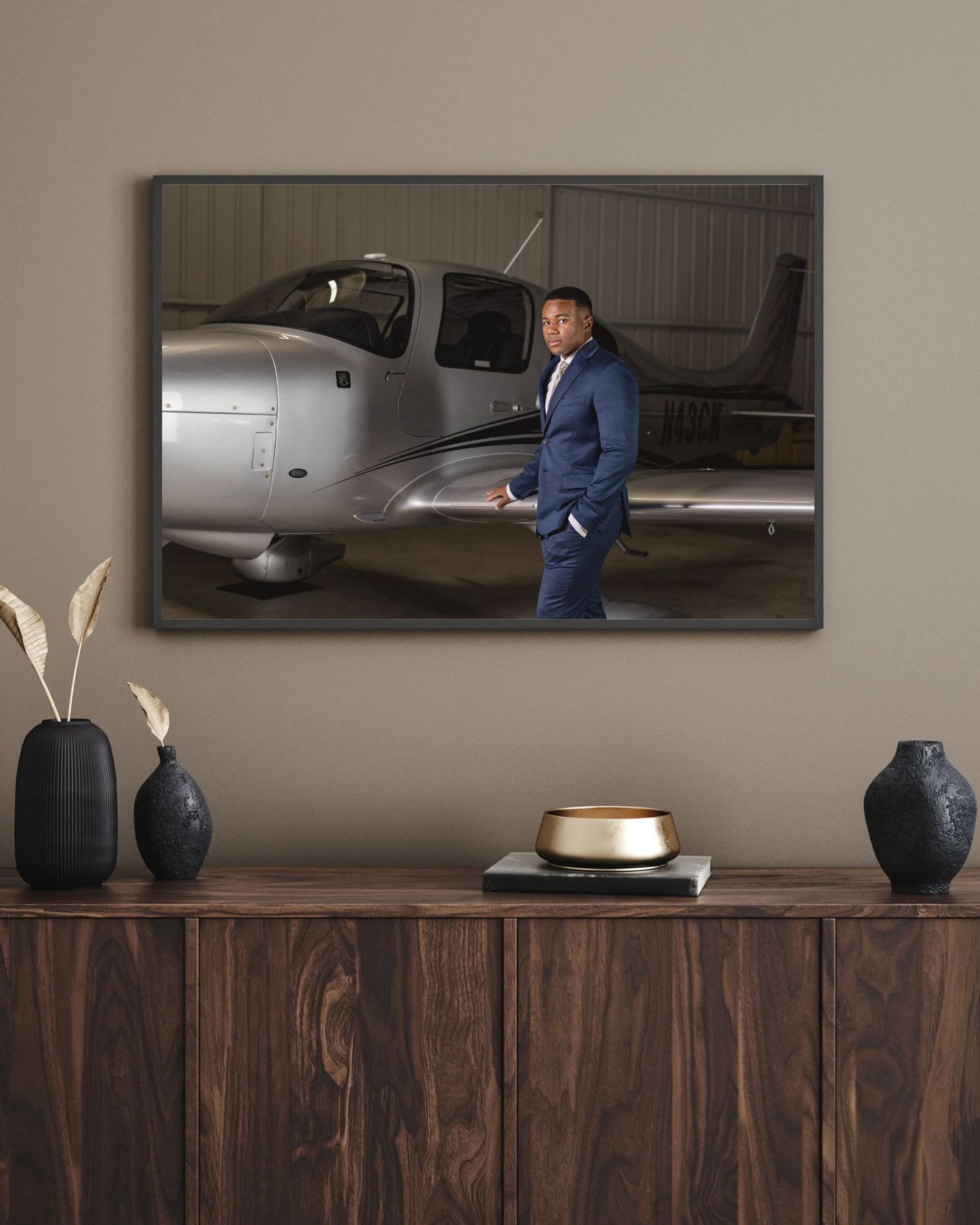 Man in navy suit standing confidently beside a silver private plane inside a hangar, framed wall art display.