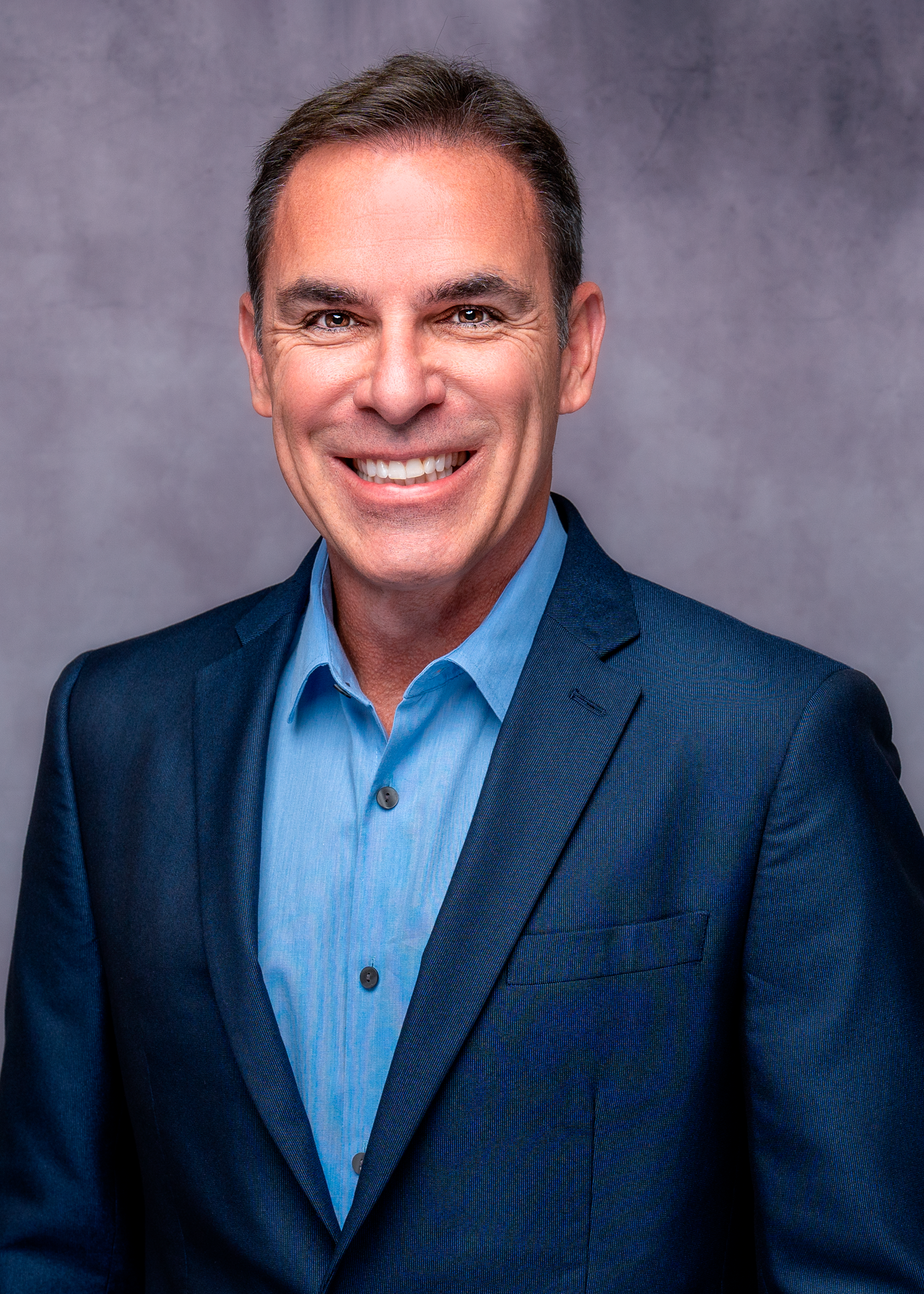 Professional headshot of a smiling middle-aged man in a navy blazer and light blue shirt against a gray background.