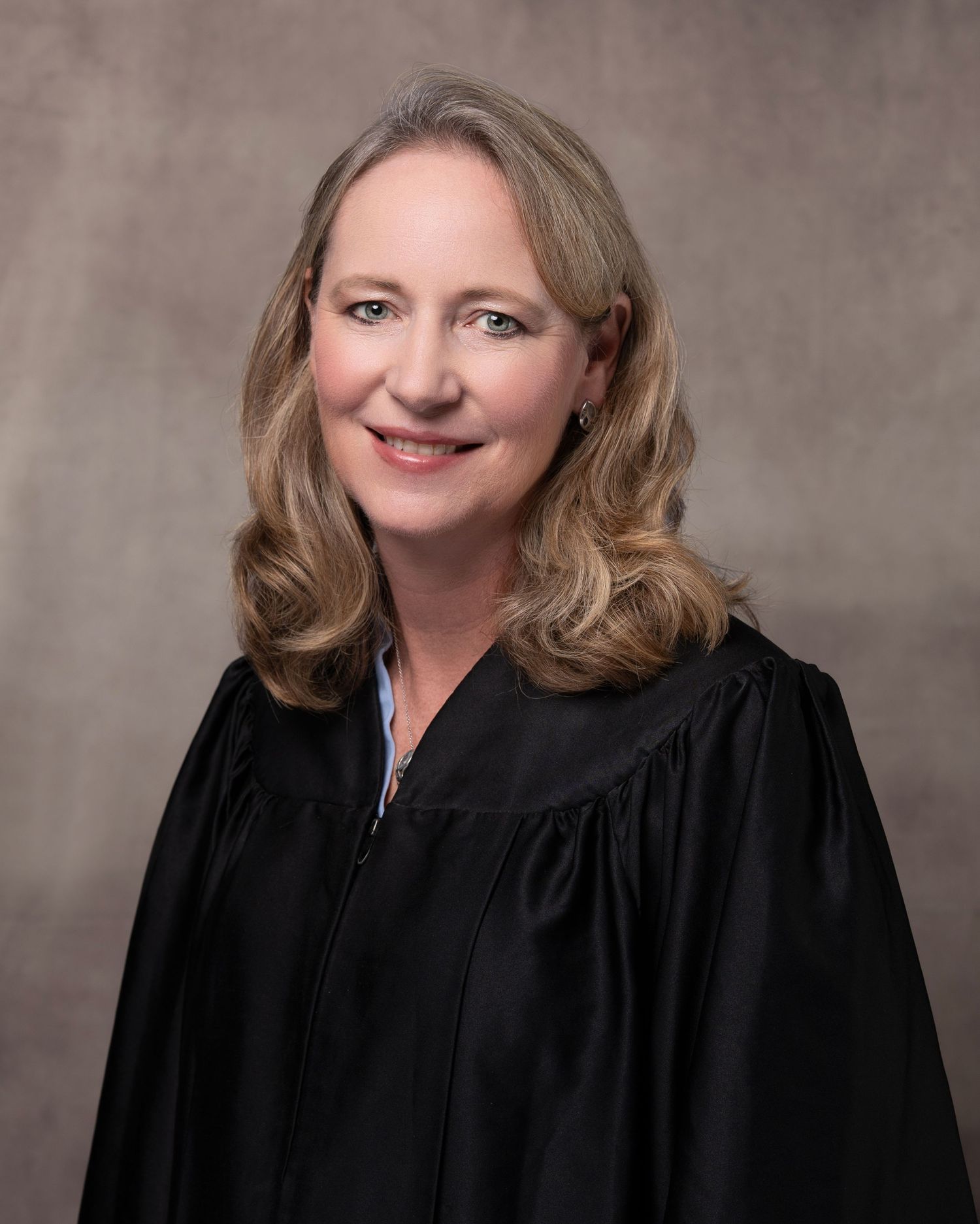 Professional portrait of a female judge wearing black judicial robes, smiling warmly against a neutral studio background.