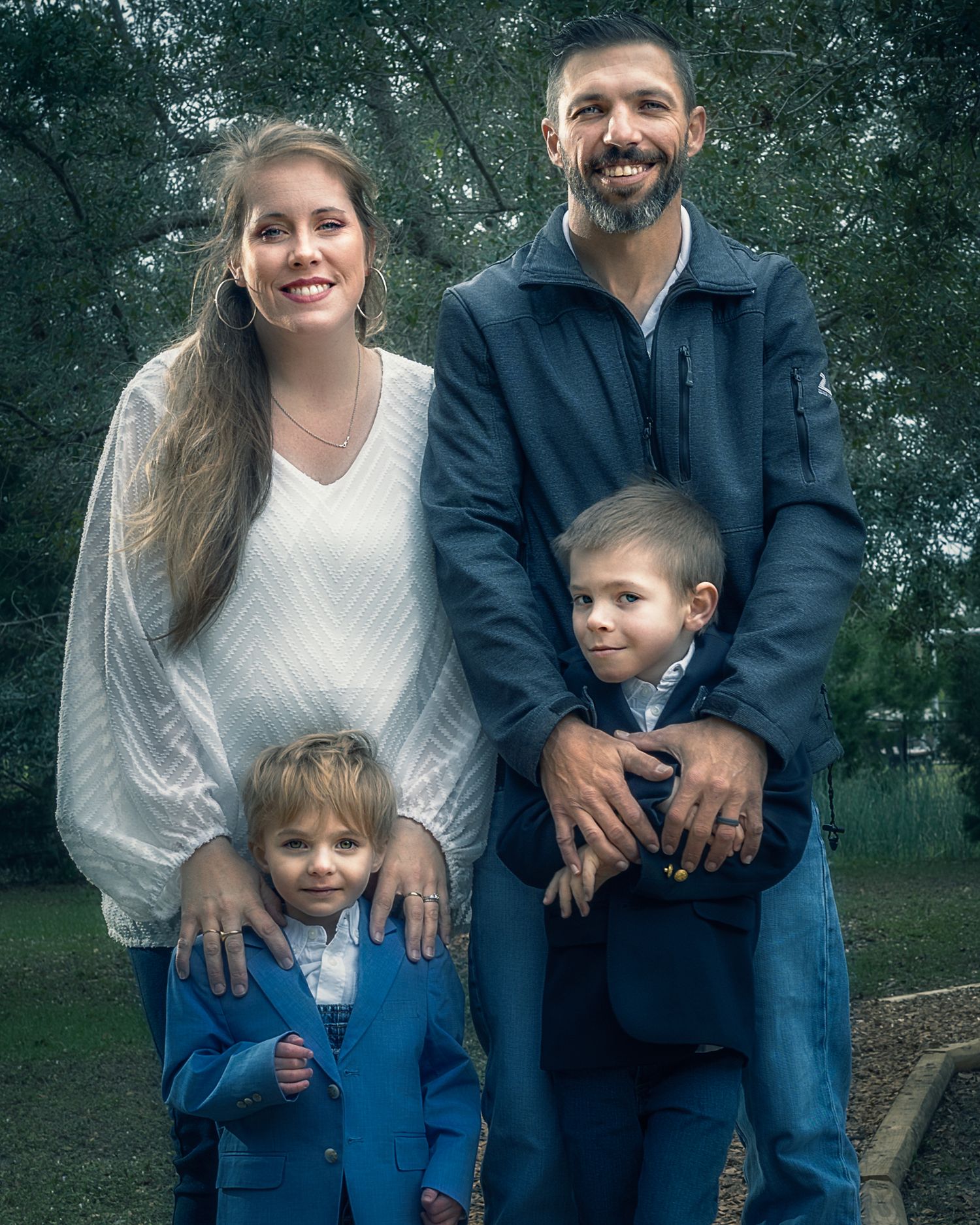 Family poses together in casual wear against a blurred outdoor background with trees and greenery.