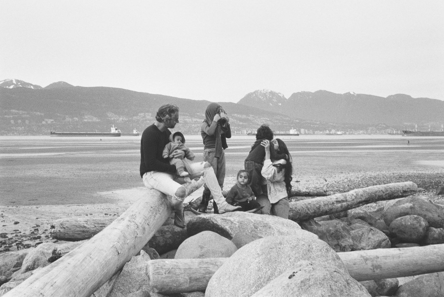 Several people sit and relax on large driftwood logs at a rocky beach with mountains visible in the background.