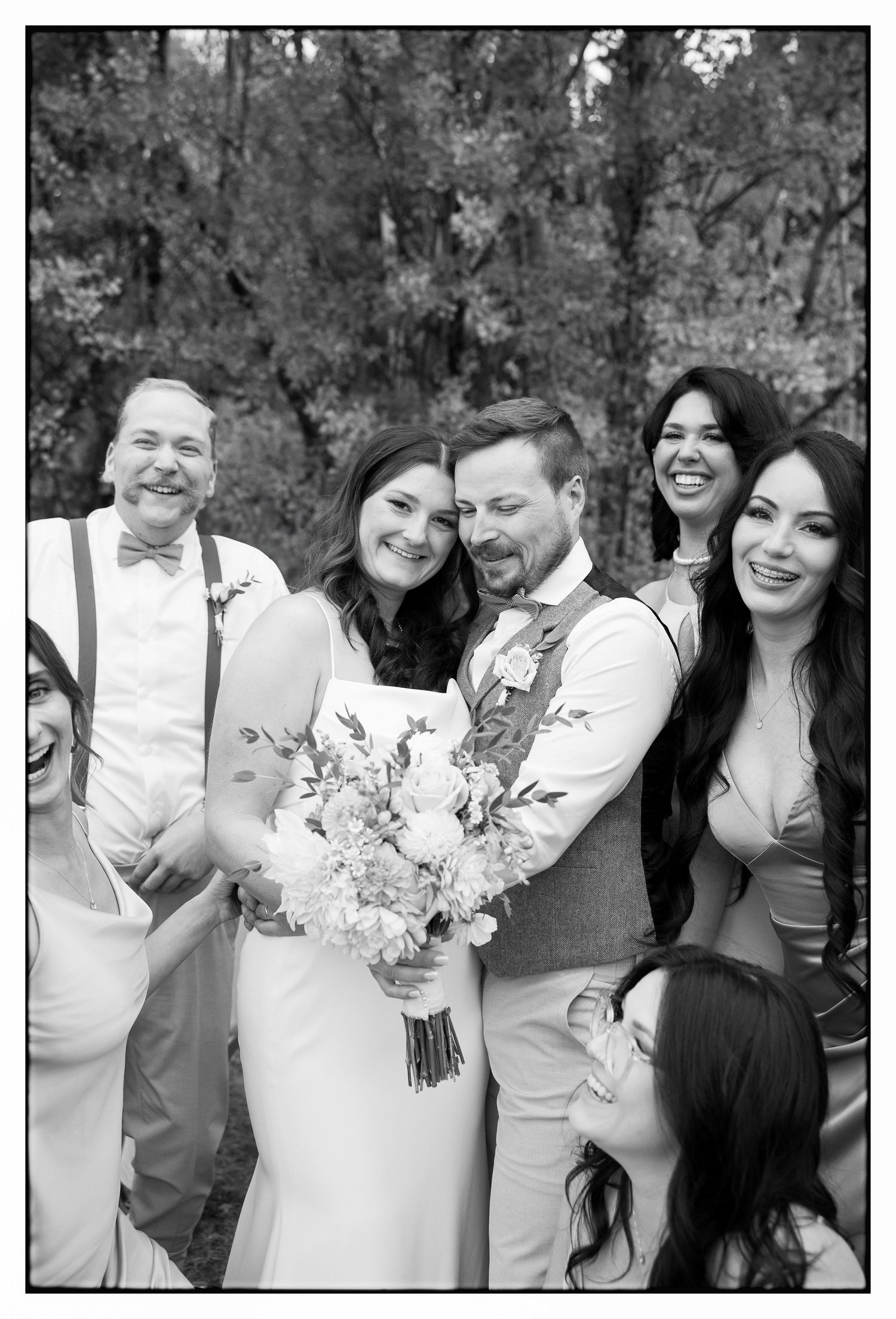A black and white wedding photo shows a happy wedding party outdoors with a floral bouquet and groomsmen in suspenders.