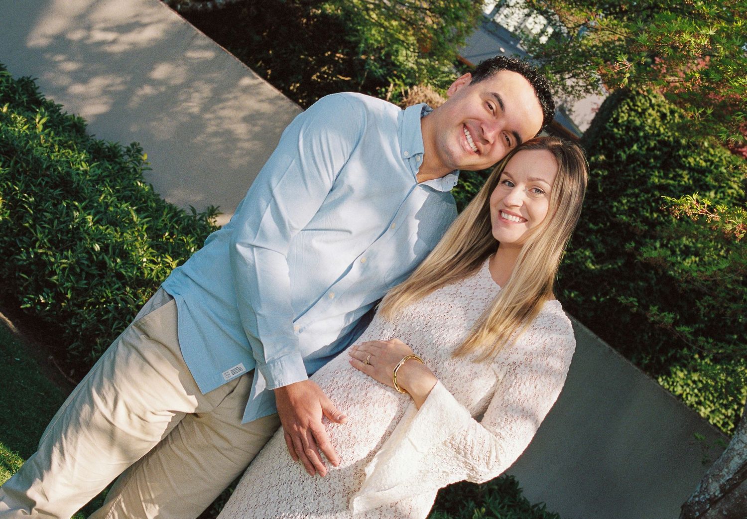 A couple in light-colored clothing smile together while posing outdoors in a garden setting.