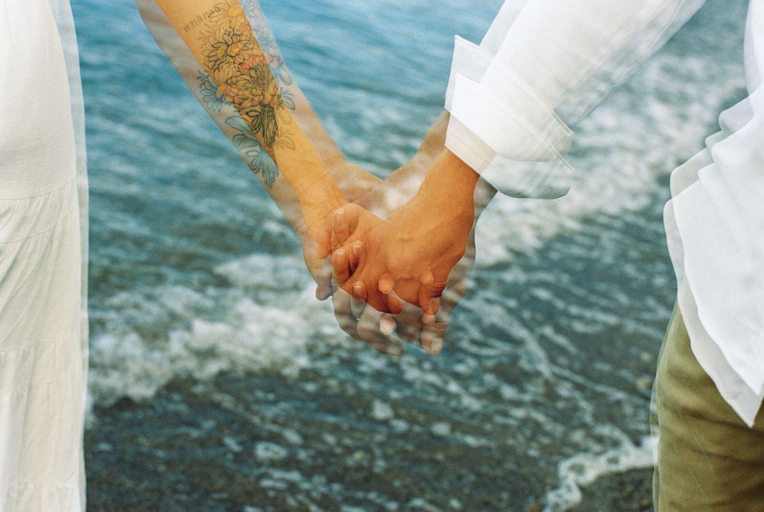 Two people in casual clothing hold hands against a backdrop of ocean waves on a sunny day by the water.