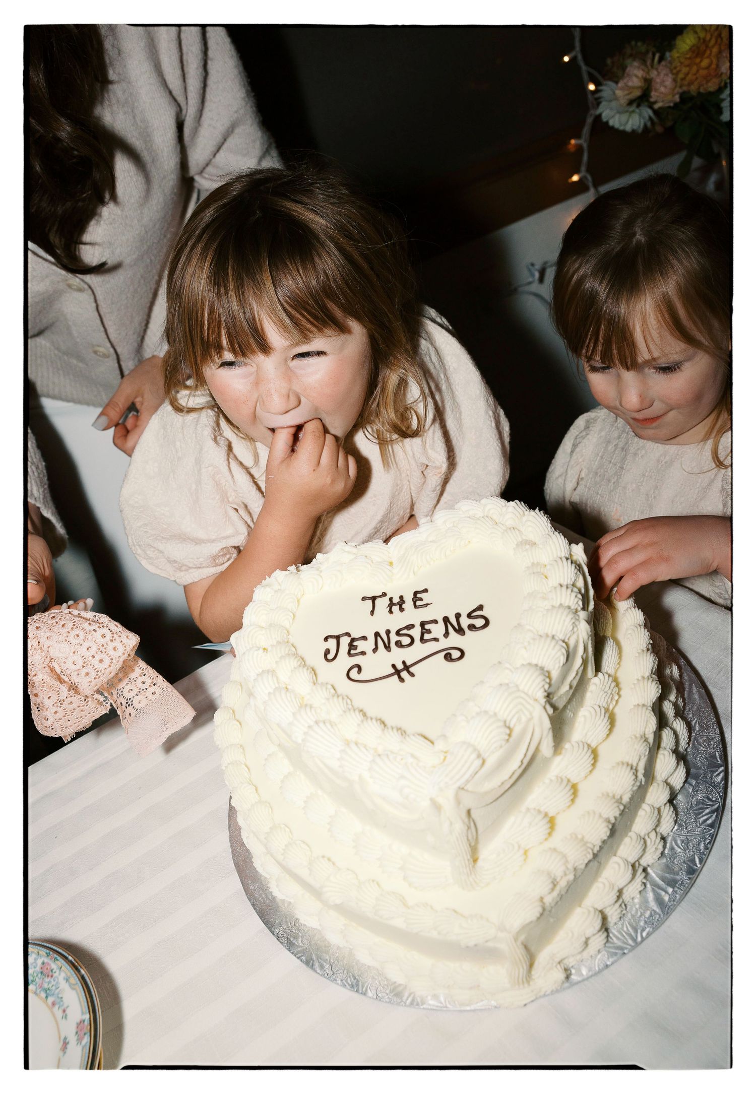 A white frosted birthday cake with 'The Jensens' written on it sits on a table while children gather around.