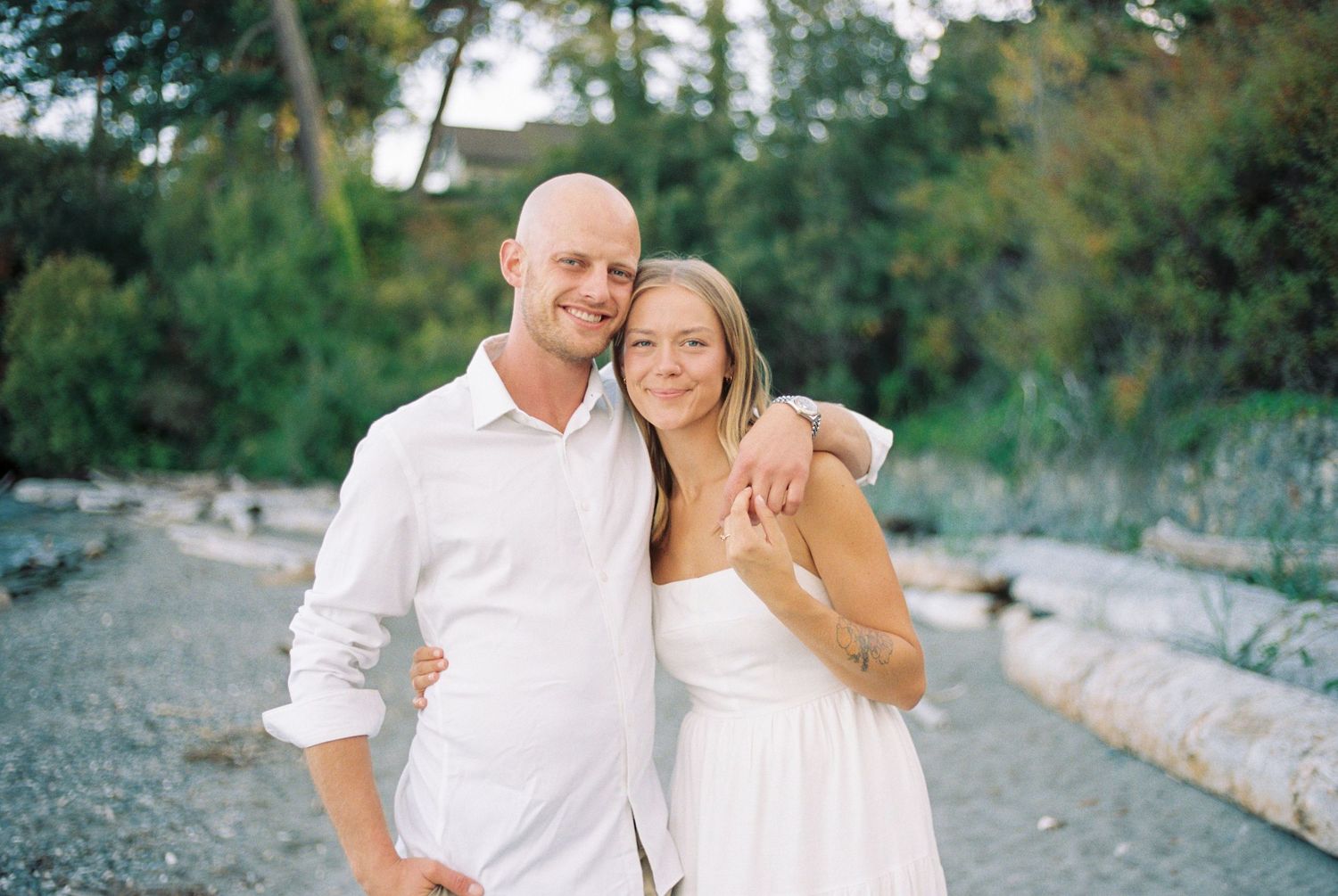 A couple in matching white outfits embraces while standing on a rocky beach path with trees in the background.