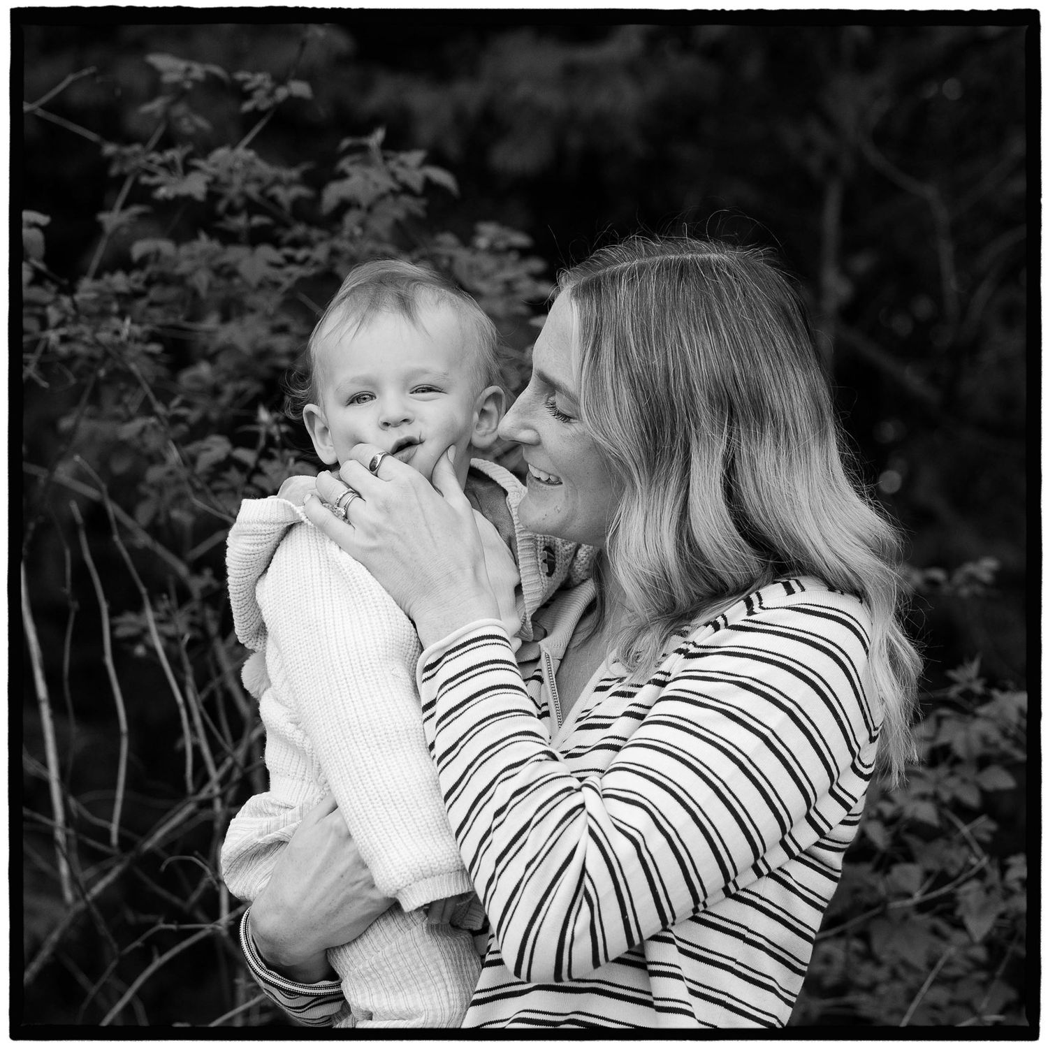 Black and white photo of a baby in the arms of someone wearing a striped shirt in an outdoor garden setting.