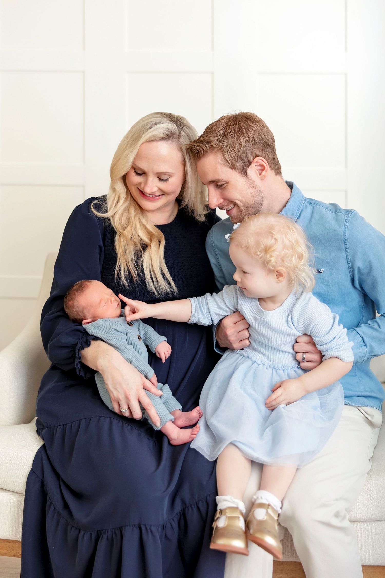 Family of four with newborn baby, toddler girl, and parents sitting together on a white couch during newborn studio session in Clear Lake IA