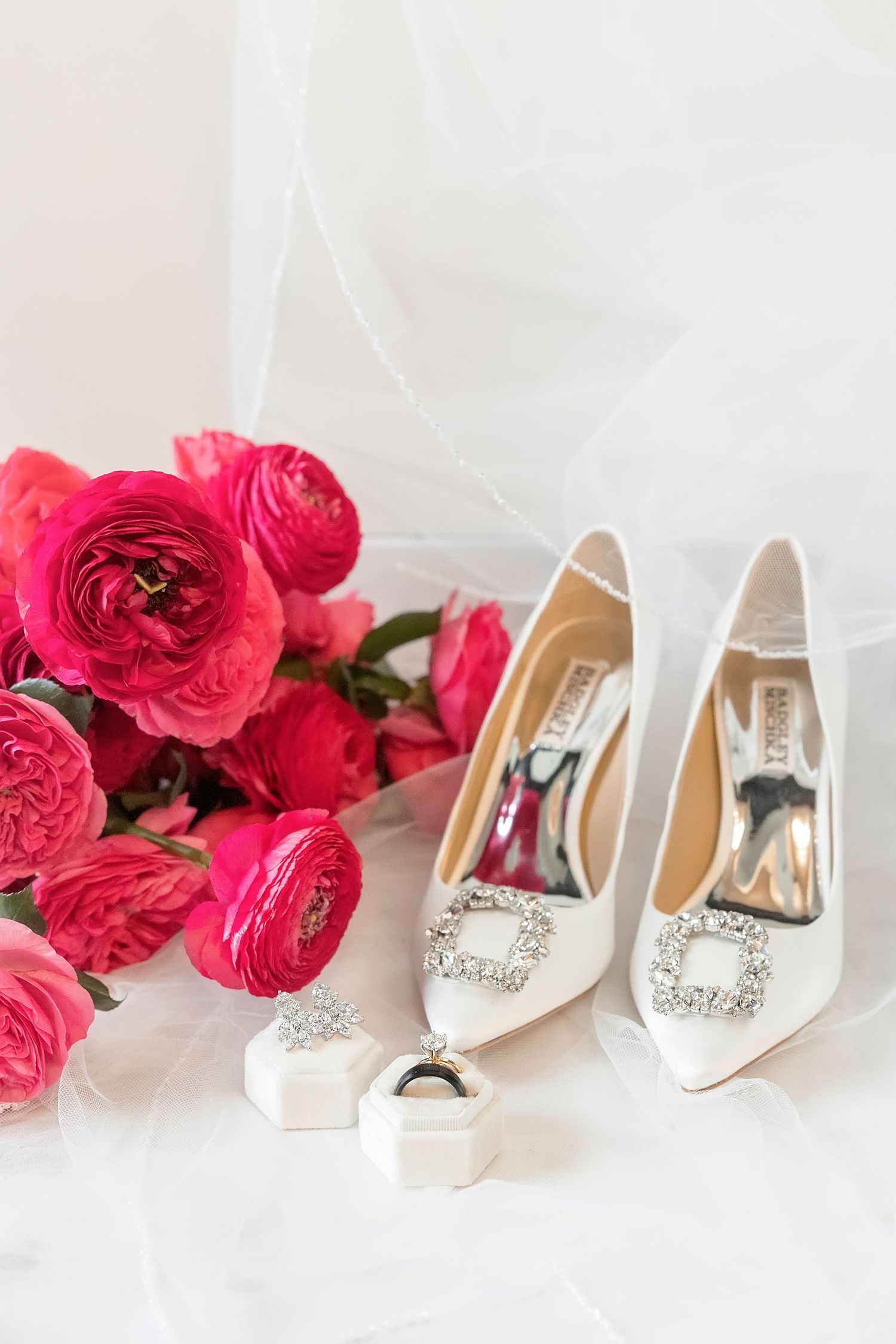 White Badgley Mischka bridal heels with crystal buckles beside pink ranunculus bouquet, diamond ring, and earrings on white veil.