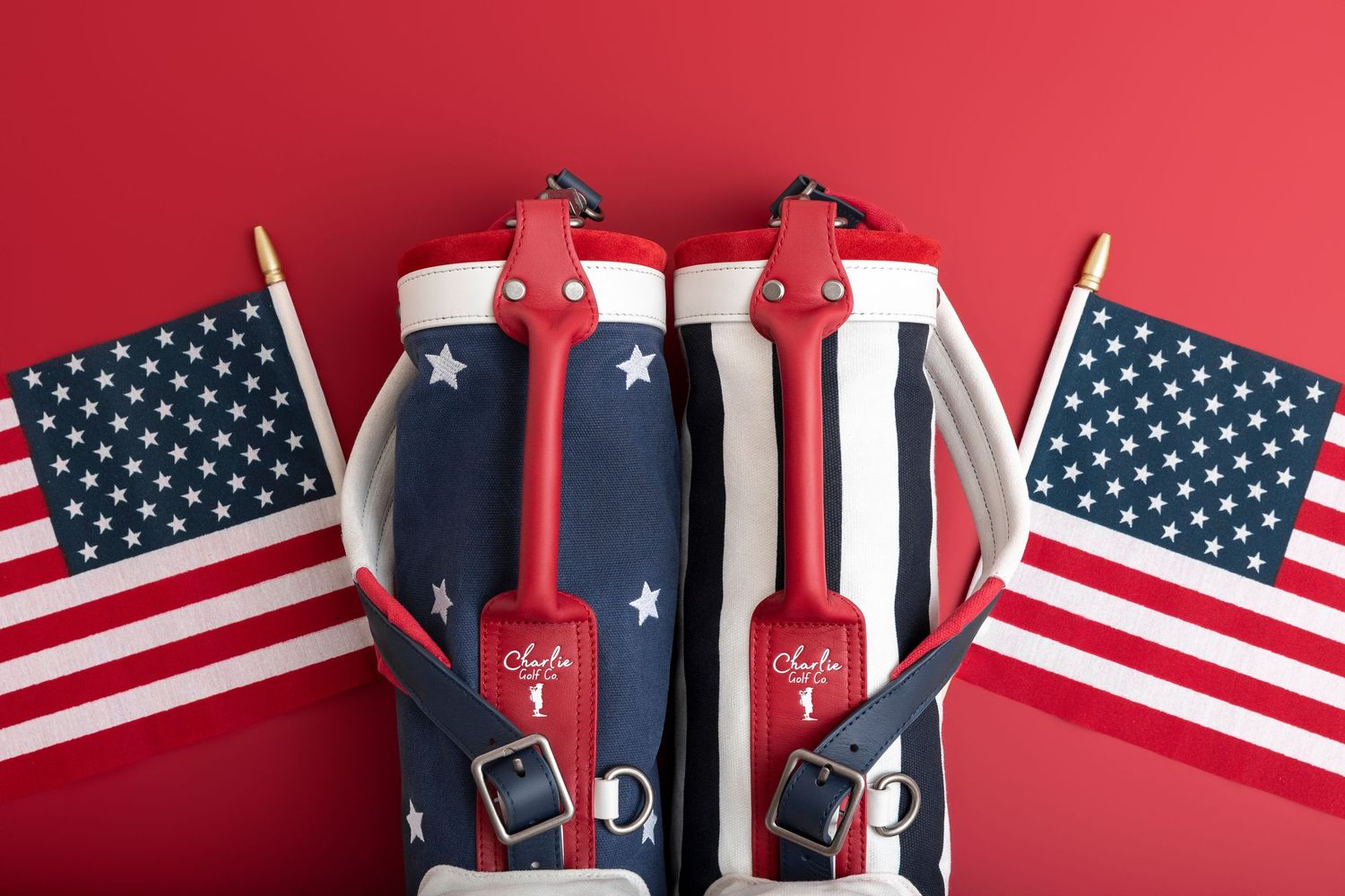 Red, white and blue stars and stripes golf bag with red leather handles and buckled straps, displayed between two small American flags.