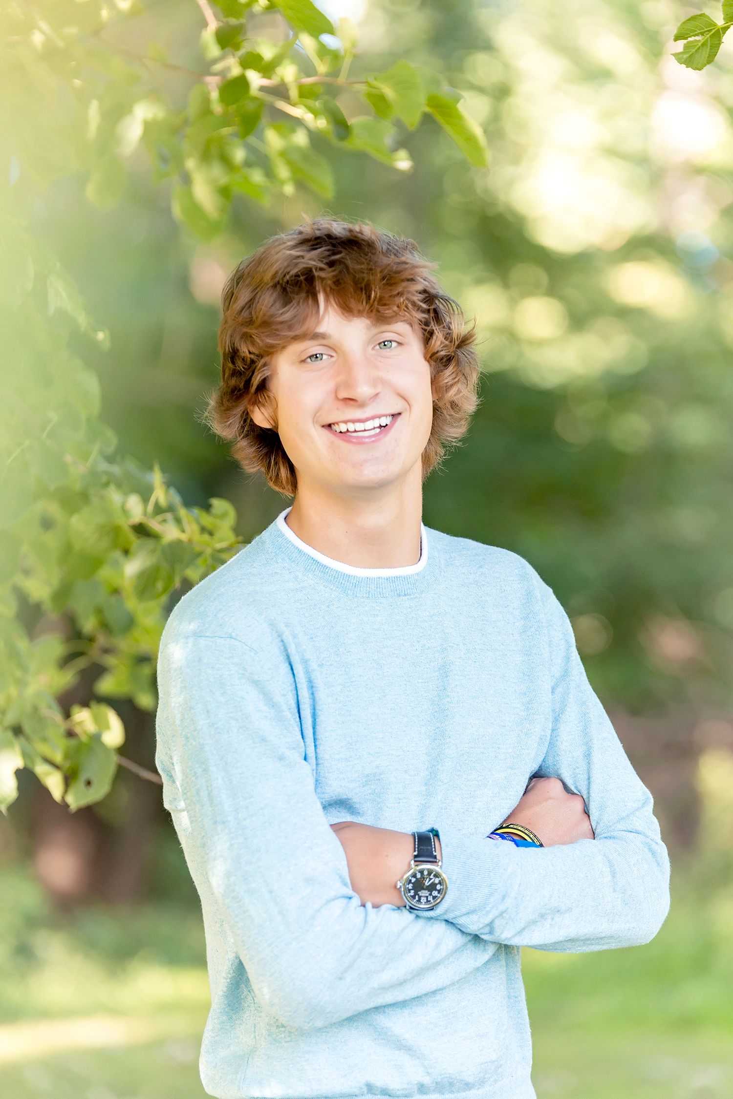 Smiling teenage boy with curly hair in light blue sweater, arms crossed, posing outdoors among green trees.