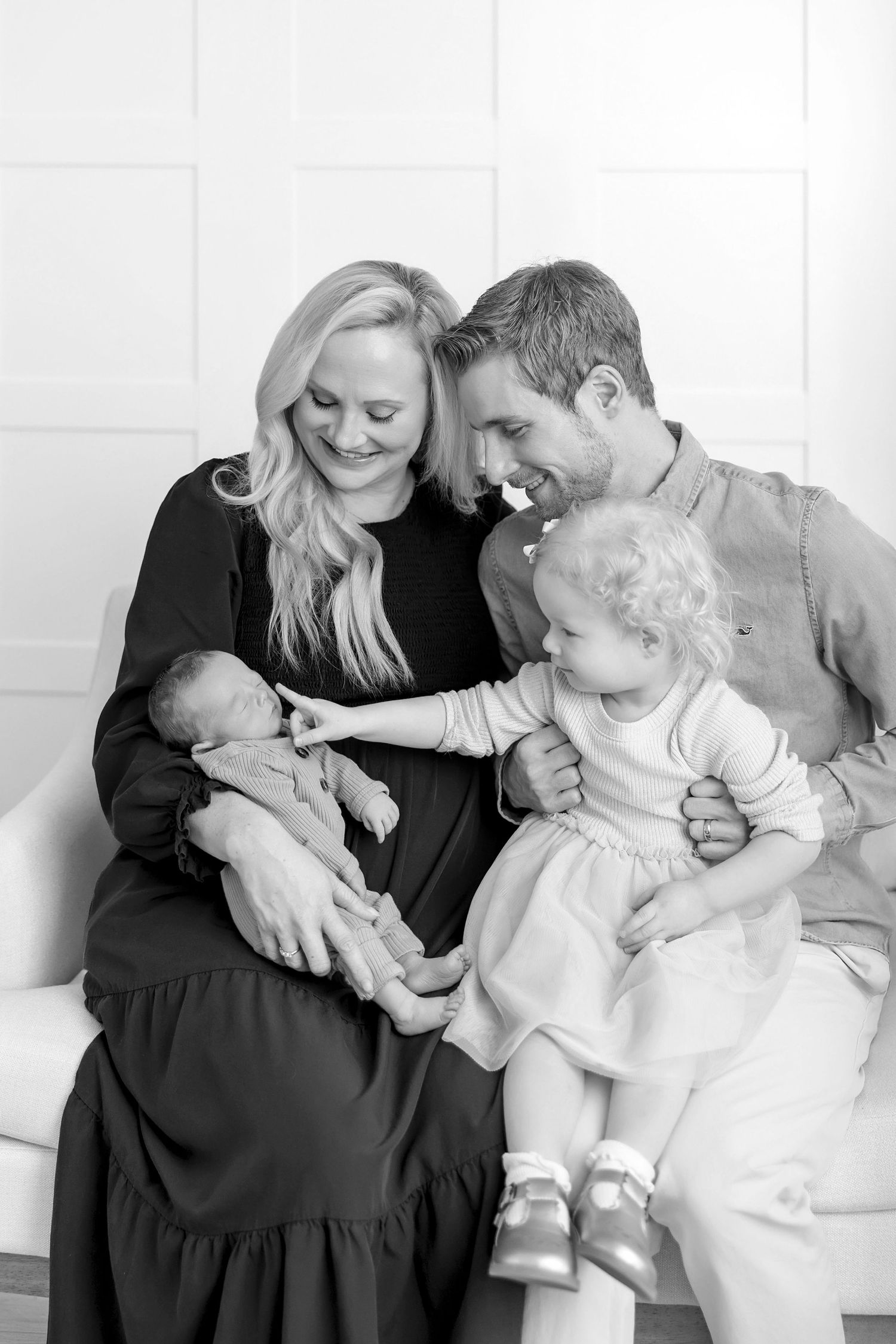 Black and white family portrait of parents with newborn baby and toddler sitting together on a white couch.