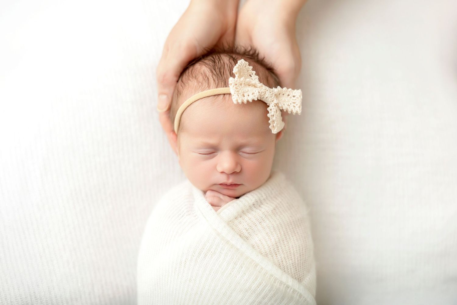 Sleeping newborn baby swaddled in white wrap with lace bow headband, cradled by gentle hands.