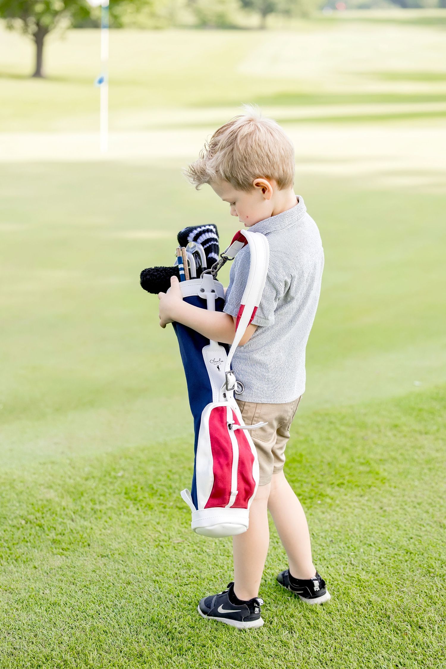 Young boy carrying a golf bag with clubs on a green golf course near the putting green.