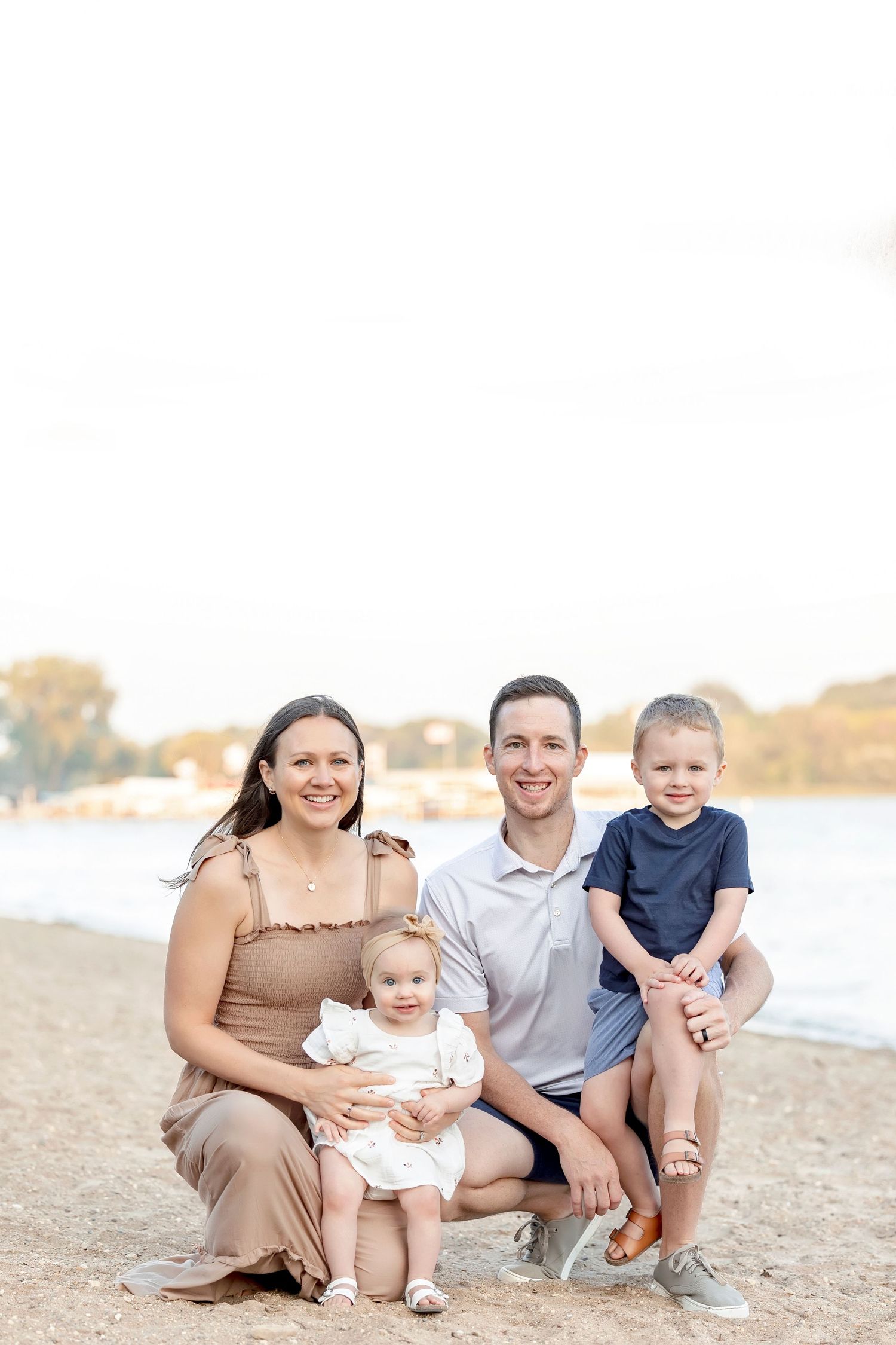 Family of four poses on sandy beach, parents smiling with toddler girl and young boy near calm water.