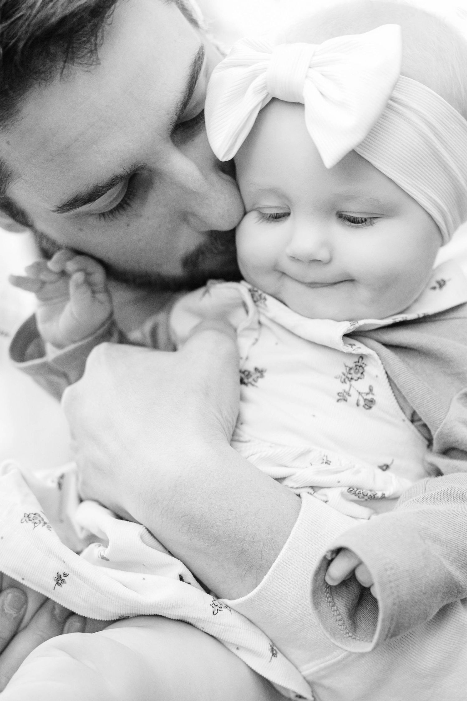 Black and white photo of a father kissing his smiling baby girl wearing a bow headband.