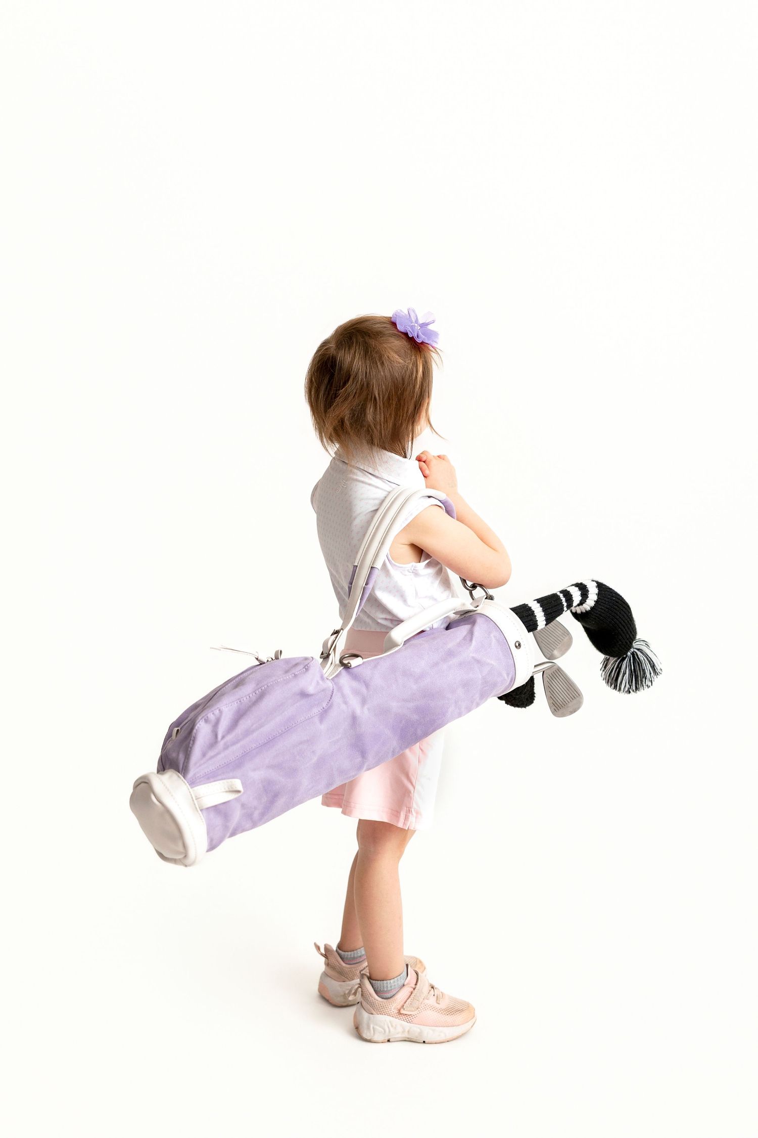Young girl carrying a purple golf bag with clubs over her shoulder, wearing a pink skirt and sneakers.