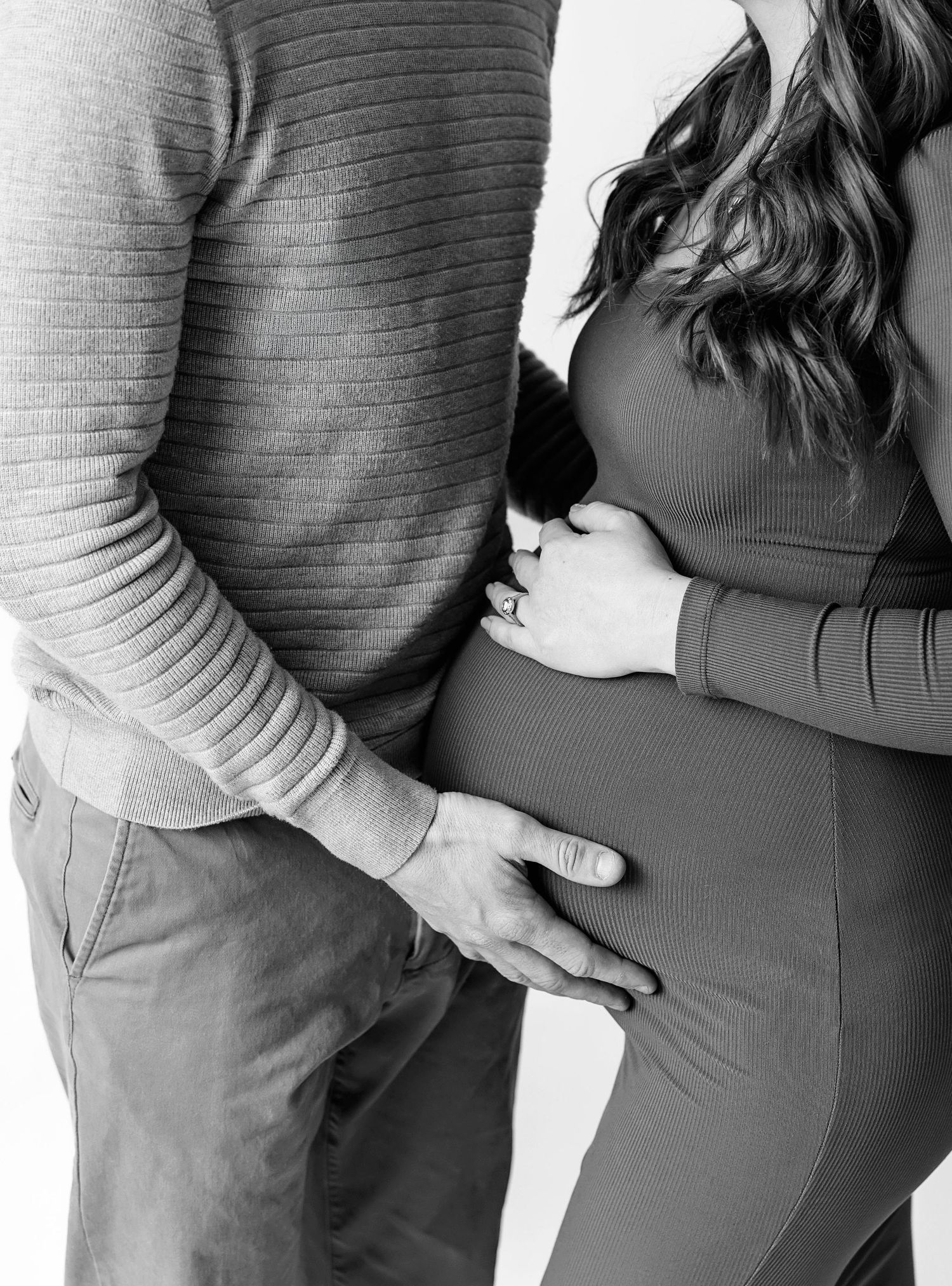 Black and white maternity photo of couple cradling pregnant belly, wearing casual knit sweater and fitted dress.