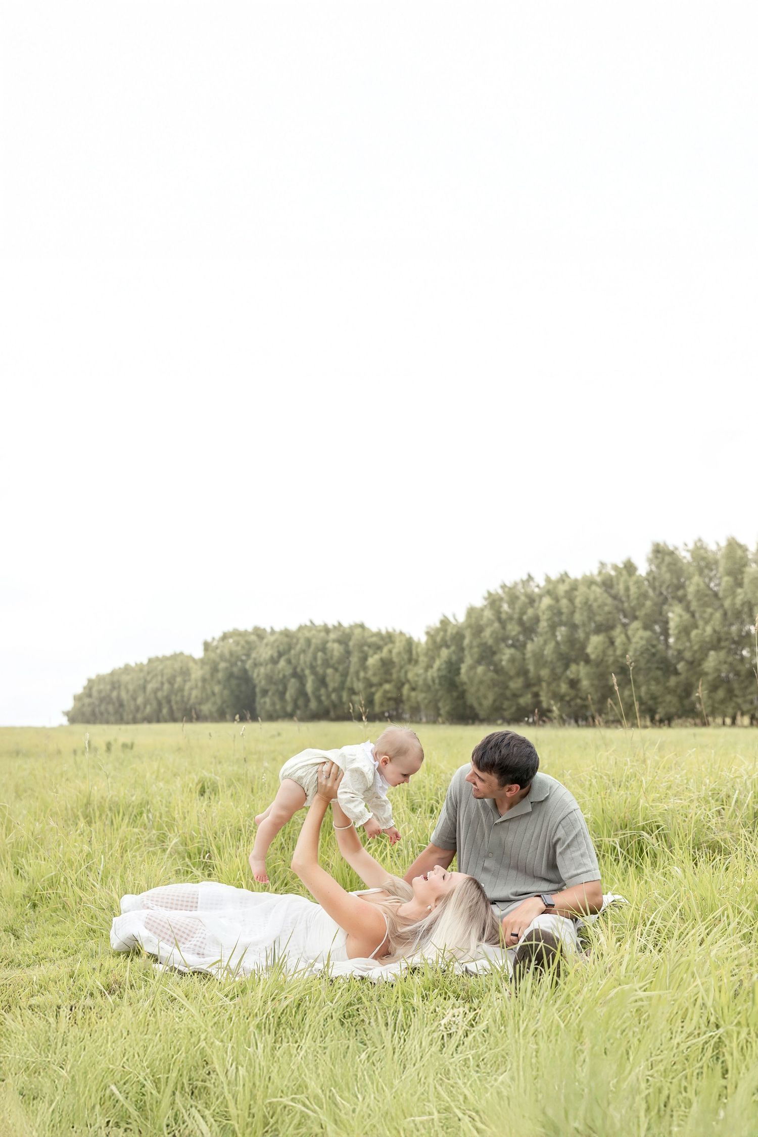 Family enjoying a sunny day in a green field, mother lifting baby while father watches lovingly.
