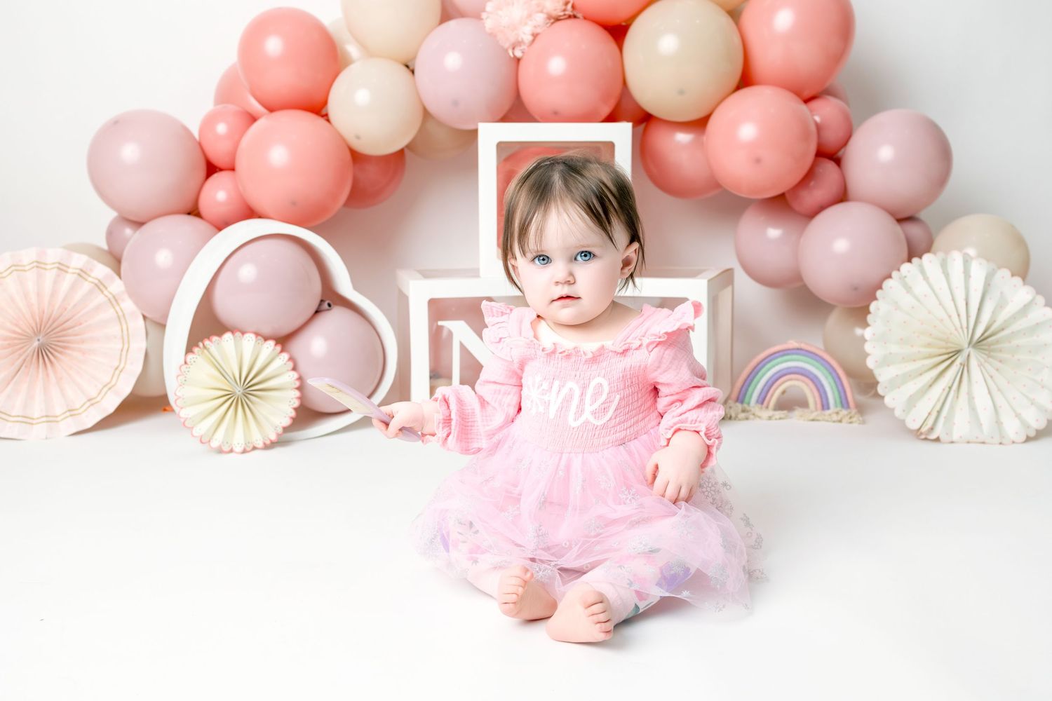 Pink and peach balloon garland with paper fan decorations against a white background creates an elegant party backdrop.