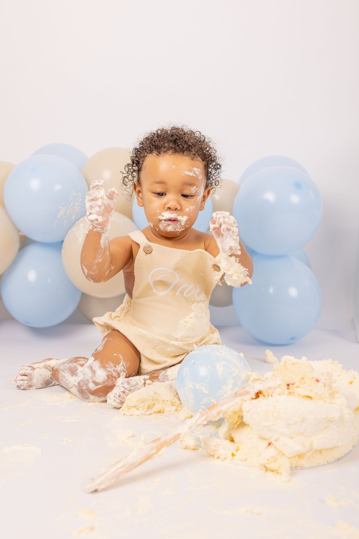 Baby in cream outfit enjoys messy cake smash photoshoot with light blue balloons and frosting everywhere.