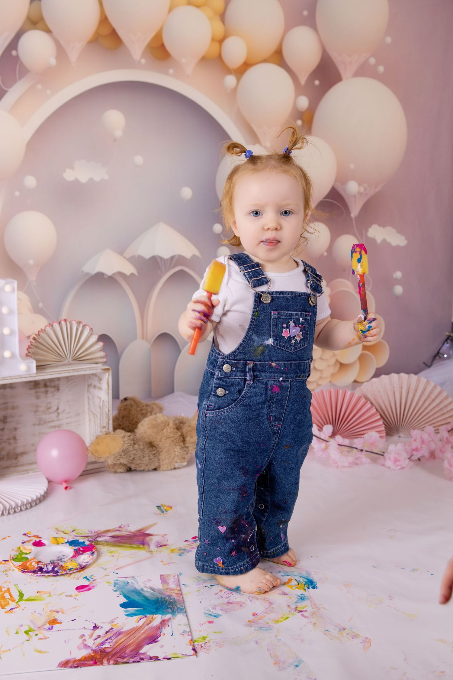 Toddler in denim overalls stands against dreamy balloon backdrop holding art supplies with colorful paint splatters on floor.