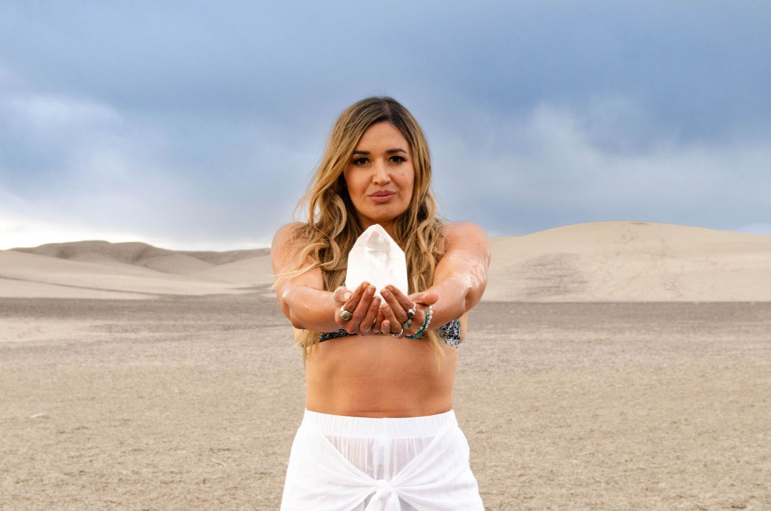 Woman in white outfit holds crystal in desert dunes under cloudy sky, showcasing spiritual healing energy.