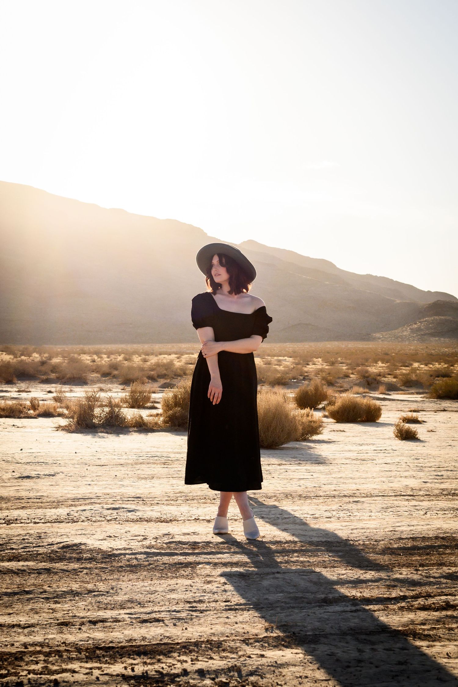 Person in a black dress twirls in the desert at sunset, casting long shadows across the arid landscape. Moody portrait Photoshoot Las Vegas.