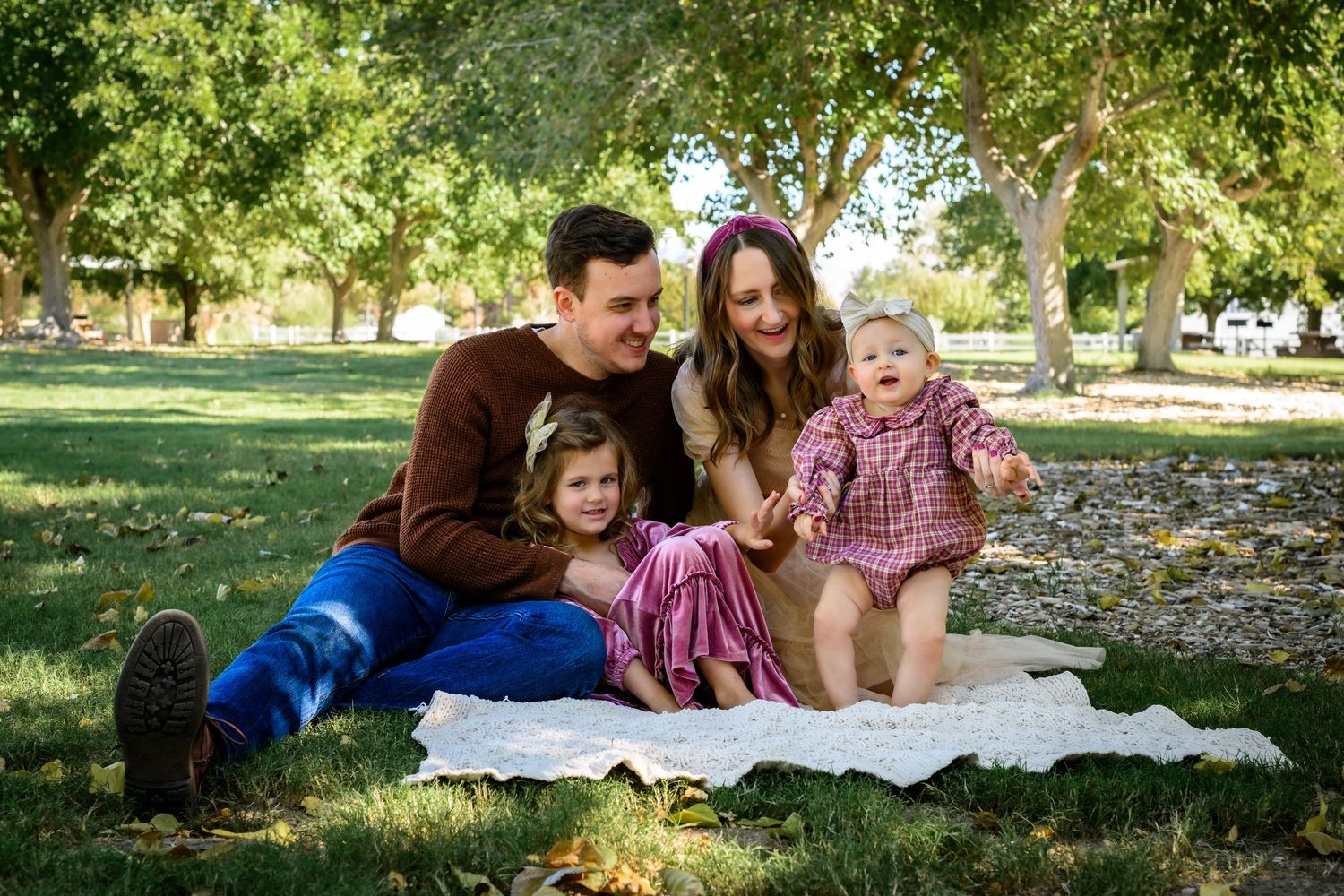 Family of four sitting on blanket in park, parents with toddler girl and baby boy smiling outdoors.