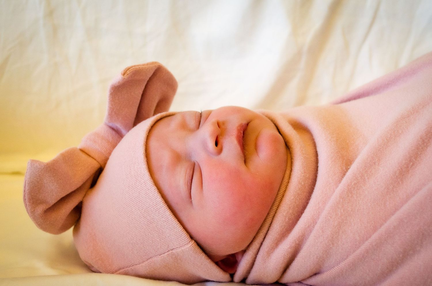 Newborn baby sleeping peacefully swaddled in pink blanket wearing pink hat with bow ears.