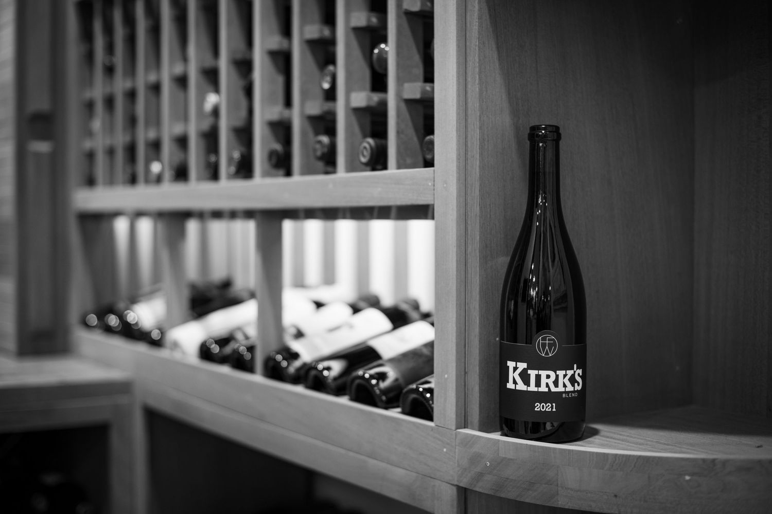Black and white photo of Kirk's wine bottle displayed on wooden wine rack shelves in a cellar.