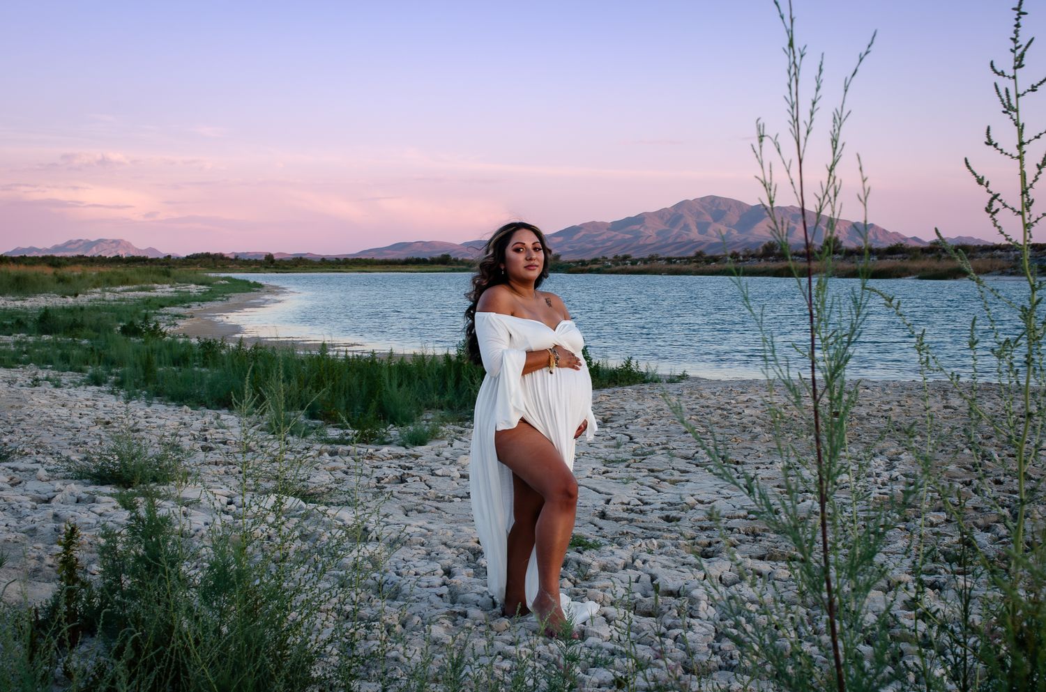 Pregnant woman in white dress poses by a lake at sunset with mountains in the background.