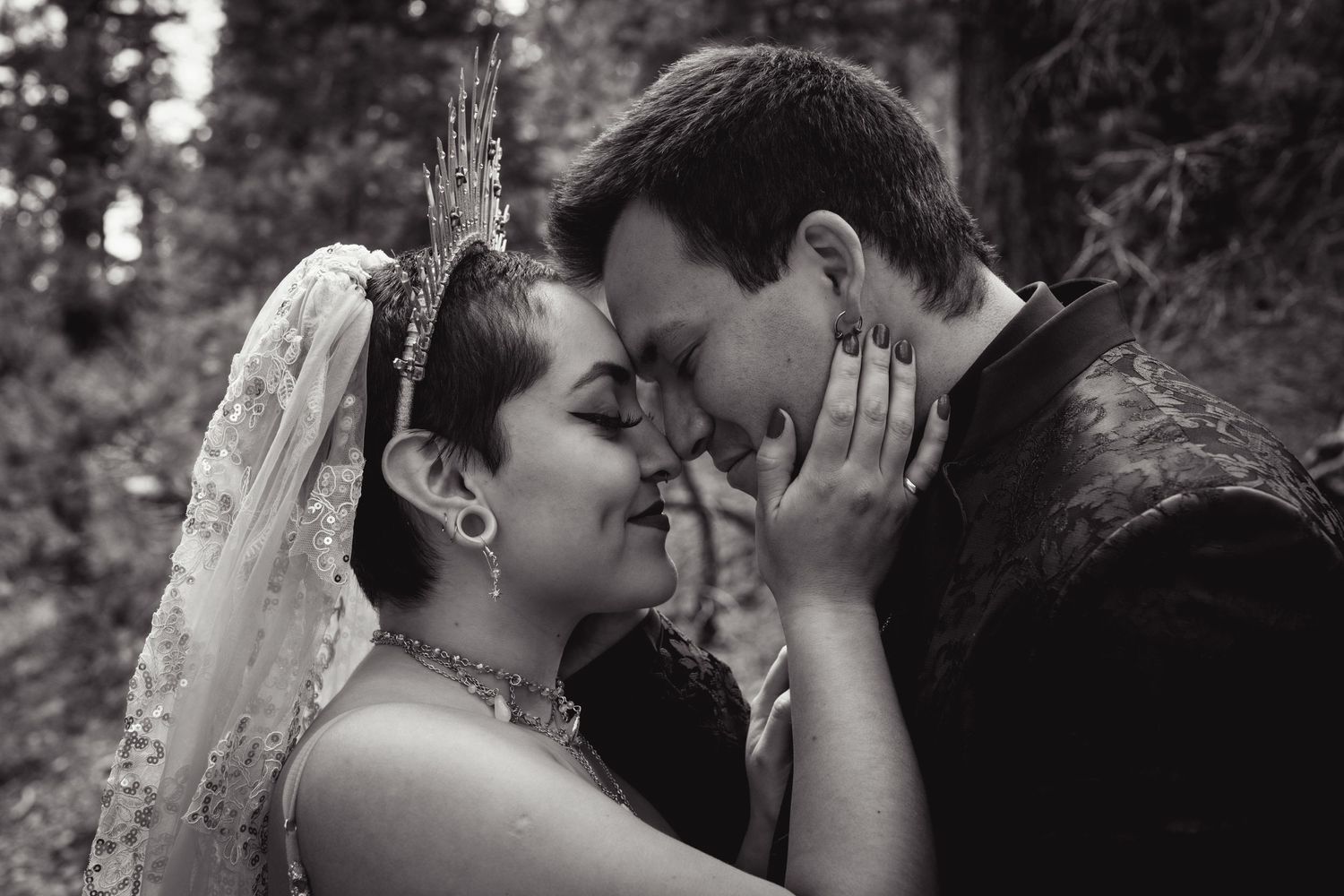 Black and white wedding photo of bride and groom touching foreheads tenderly outdoors among trees.