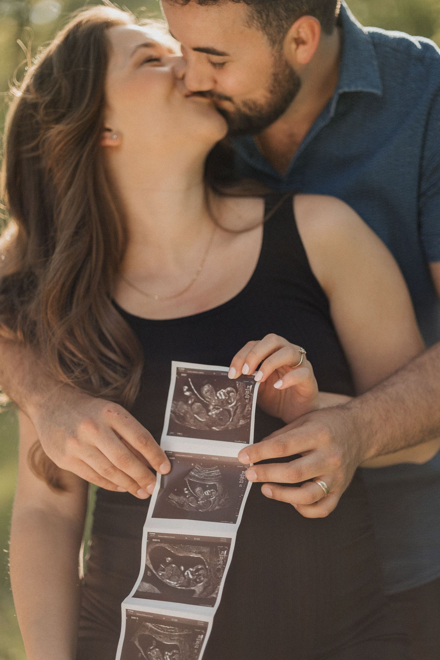 An expecting couple kisses while holding ultrasound photos in a warm, sun-drenched outdoor setting.